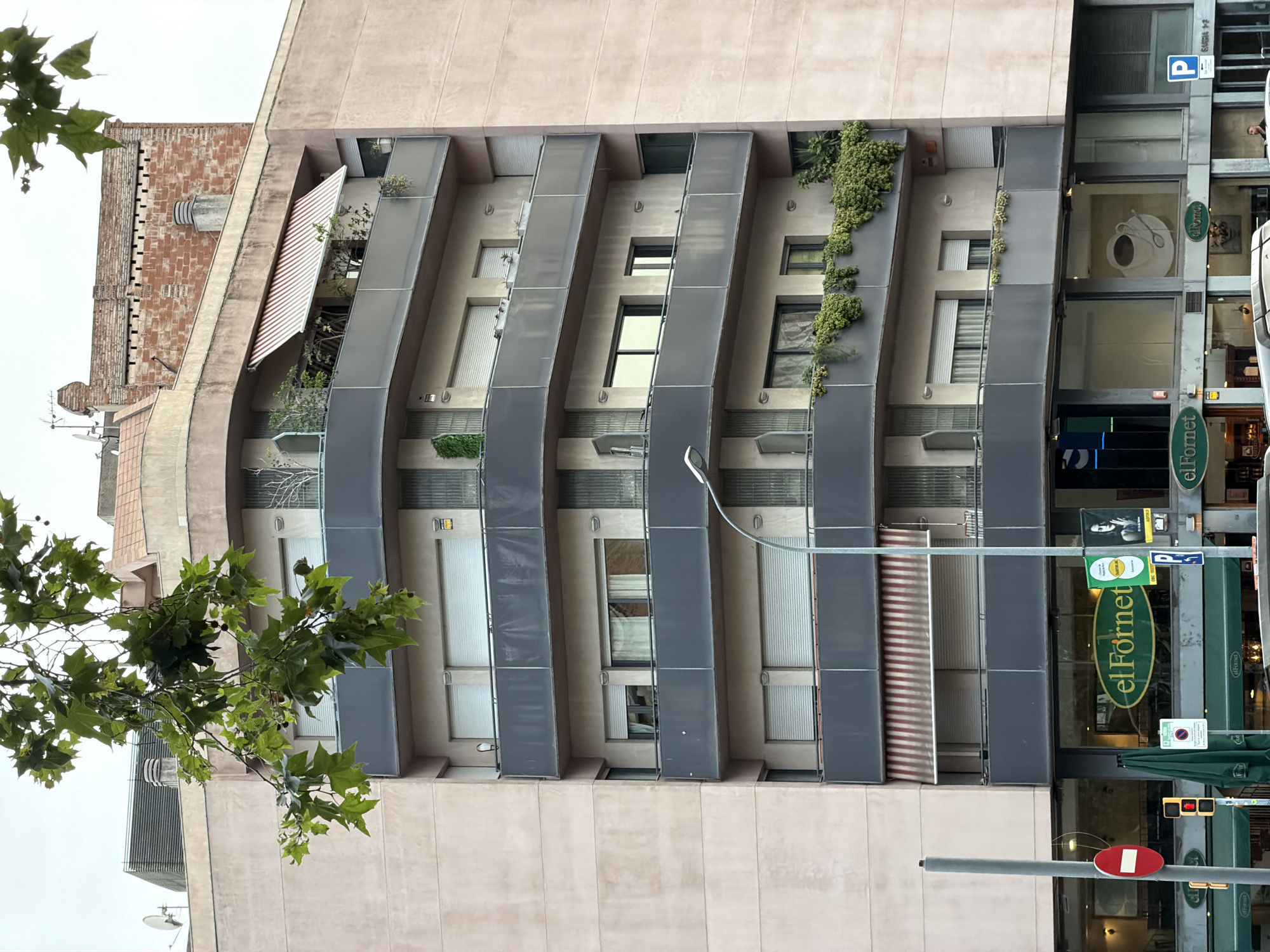 The image depicts a street-level view of a multi-story building in Barcelona, Spain, with a prominent "El Fornet" bakery sign visible. The building features multiple balconies with dark grey railings, some adorned with greenery. Various windows and awnings are visible on the facade. In the foreground, traffic lights, a red circular no-entry sign, and a light pole are present. The sky is overcast. The overall impression is a typical urban street scene.
