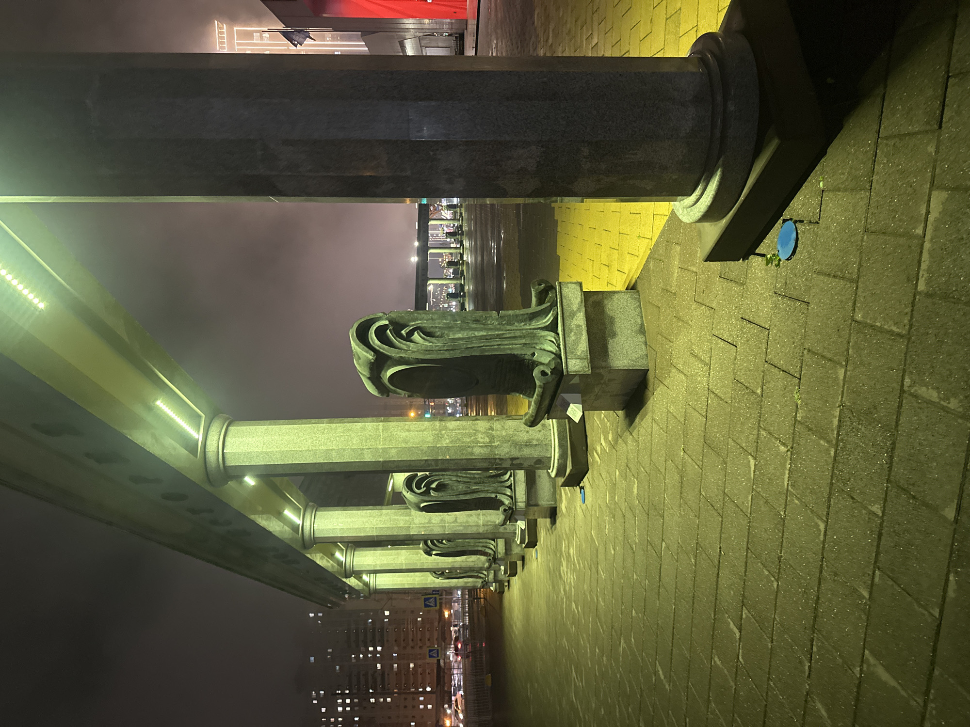 The image depicts a colonnade at night, likely in Stavropol, Russia. The foreground features a row of stone pillars, topped with decorative capitals. A low, illuminated roof structure runs above the columns. To the right, a paved walkway extends into the distance, illuminated by yellow lighting. Beyond the colonnade, there is a dark body of water and a cityscape with illuminated buildings. The sky is overcast, obscuring the details of the skyline. There are no people visible in the image. A small blue circle is affixed to the walkway near a column.