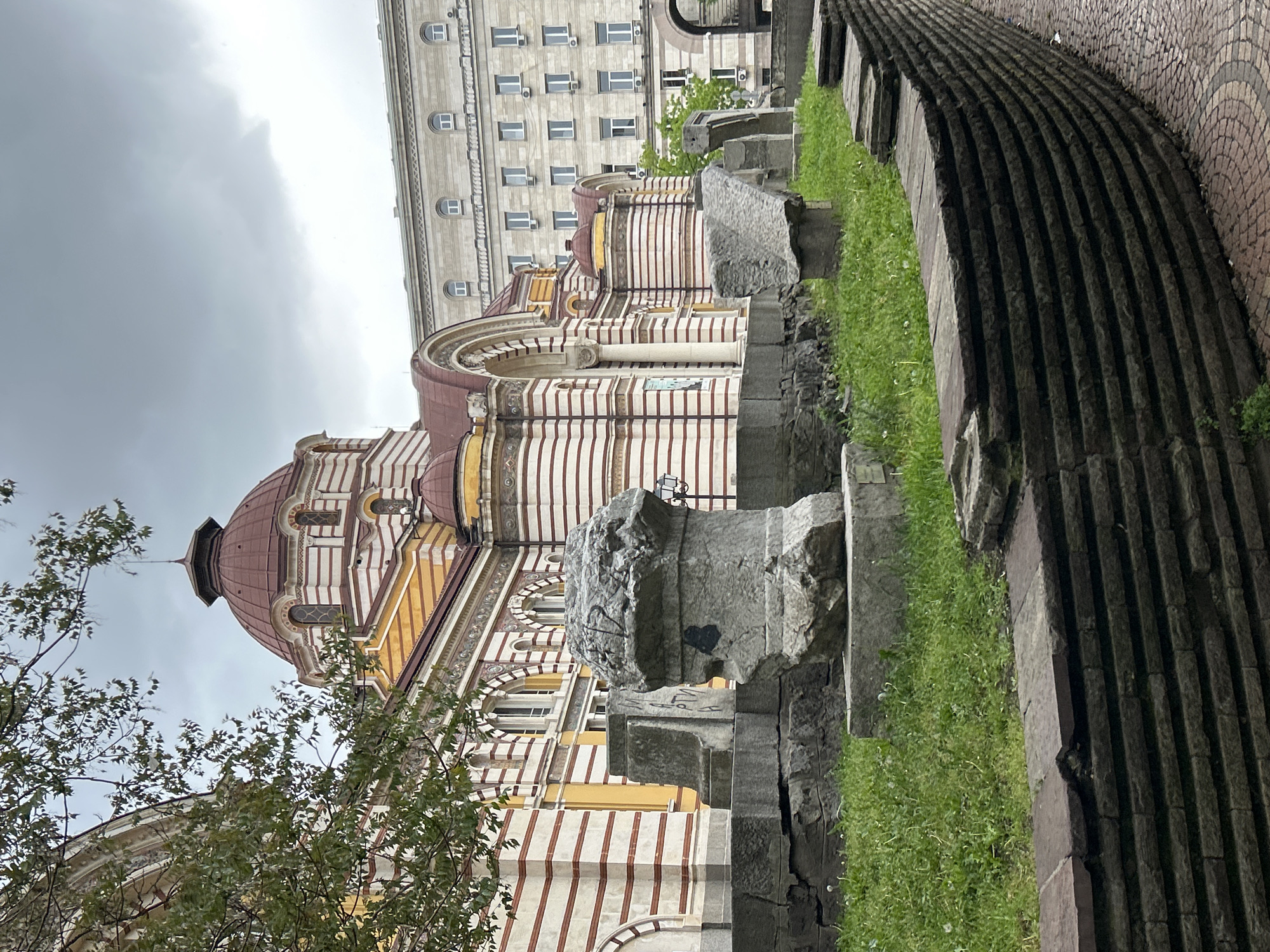 This is a ground-level, medium-shot photograph taken in Sofia, Bulgaria, featuring the Church of St. George behind what appears to be a remnant of Roman-era walls and columns.
The sky is overcast and the foreground is defined by the ruins with grass growing between the stones. The Church of St. George has a distinctive striped facade in cream and red brick with a large dome. In the background, there is a building with multiple windows, suggestive of a government or apartment building in the city center. The image is rotated 90 degrees.