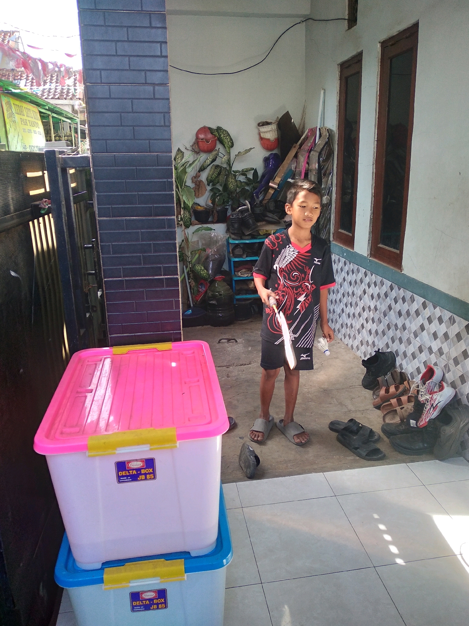 A young boy stands outside a dwelling in Cimahi, Indonesia, holding a badminton racket and shuttlecock. He wears a black t-shirt with red and white accents and black shorts. The boy stands on a tiled floor, with several pairs of sandals and shoes scattered nearby. Behind him, there are two large plastic storage bins, the top one pink and the bottom one blue. Further back, a dark brick column is visible, along with windows and what appears to be storage shelves filled with various items. The environment suggests a residential outdoor area.