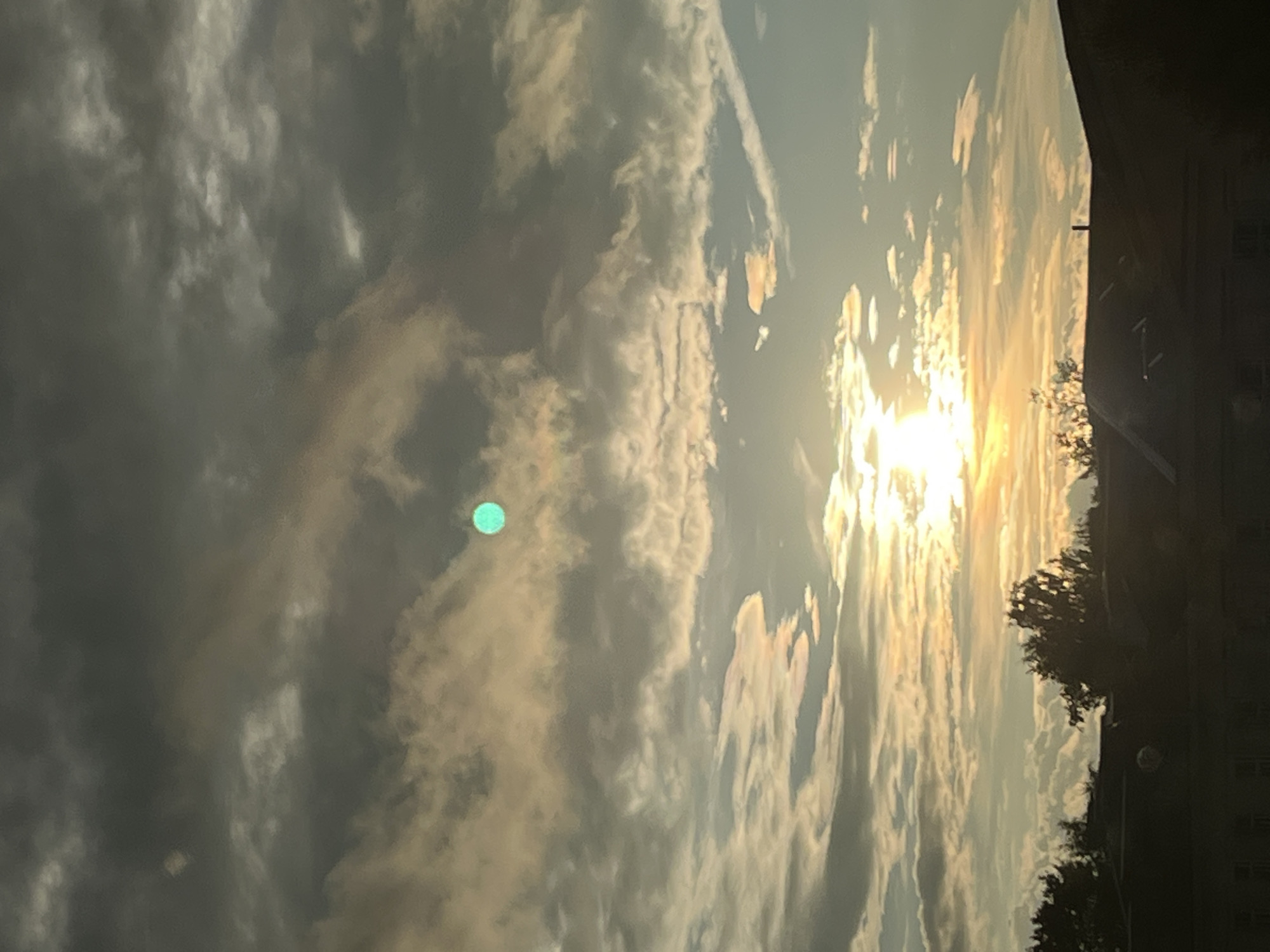 The image captures a sky view above the city of Kohtla-Järve, Estonia, during what appears to be sunset or sunrise. Dark, heavy clouds dominate the upper portion of the frame, with patches of lighter, illuminated clouds in the middle and lower sections, suggesting the sun is low on the horizon and breaking through. A distinct, bright, teal-colored circular object is visible in the upper middle section of the sky, amidst the clouds. The sun itself is a bright, horizontal band of light near the center of the image, casting a warm glow. Silhouetted against the bright sky are the dark shapes of trees and the rooftops of buildings, with some faint details of windows visible on the buildings in the foreground. The overall impression is one of a dramatic sky with diffused sunlight and atmospheric conditions.
