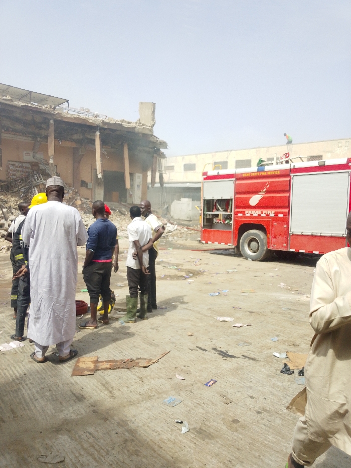 The scene is located outdoors in Fagge C, Nigeria, depicting the aftermath of a structural incident. In the left background, a two-story building is partially collapsed, with visible charring and significant structural damage to its upper floor and roof. Faint smoke emanates from the damaged structure.
In the mid-ground, a red fire truck, labeled "KANO STATE FIRE SERVICE" and "PRIME FIRE," is parked. A person in a green/yellow shirt stands on a ladder on the fire truck's roof. Another individual in a green/yellow shirt is visible on the roof of an adjacent, less damaged building in the far background.
In the foreground and mid-ground, a group of approximately six men are gathered, facing towards the damaged building. One man wears a white traditional garment and cap. Another individual wears a yellow hard hat and a dark uniform, consistent with a firefighter. Other men in the group wear casual attire, including one in a blue shirt and another in a white shirt and green rubber boots. In the far foreground right, a partially visible individual wears a light-colored traditional garment.
The ground consists of concrete, scattered with debris including cardboard, plastic wrappers, and various litter, alongside dark liquid stains. The sky is clear.
