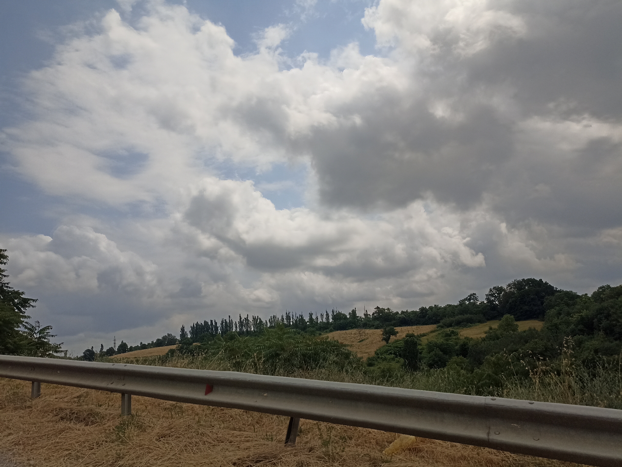 The image depicts a rural landscape with a guardrail in the foreground. The guardrail, made of metal, runs horizontally across the frame, appearing to be a safety barrier on the side of a road. Beyond the guardrail, the landscape unfolds into rolling hills covered with dry, golden grass. Further in the distance, there is a dense line of trees that forms a dark green horizon. Above the trees, a dramatic sky is filled with large, fluffy white and grey clouds, suggesting an overcast or potentially stormy day. The scene is consistent with a view from a road in a rural area, possibly during the summer months given the dry vegetation. There are no people or animals visible in the image. The location is identified as Digah, Azerbaijan.