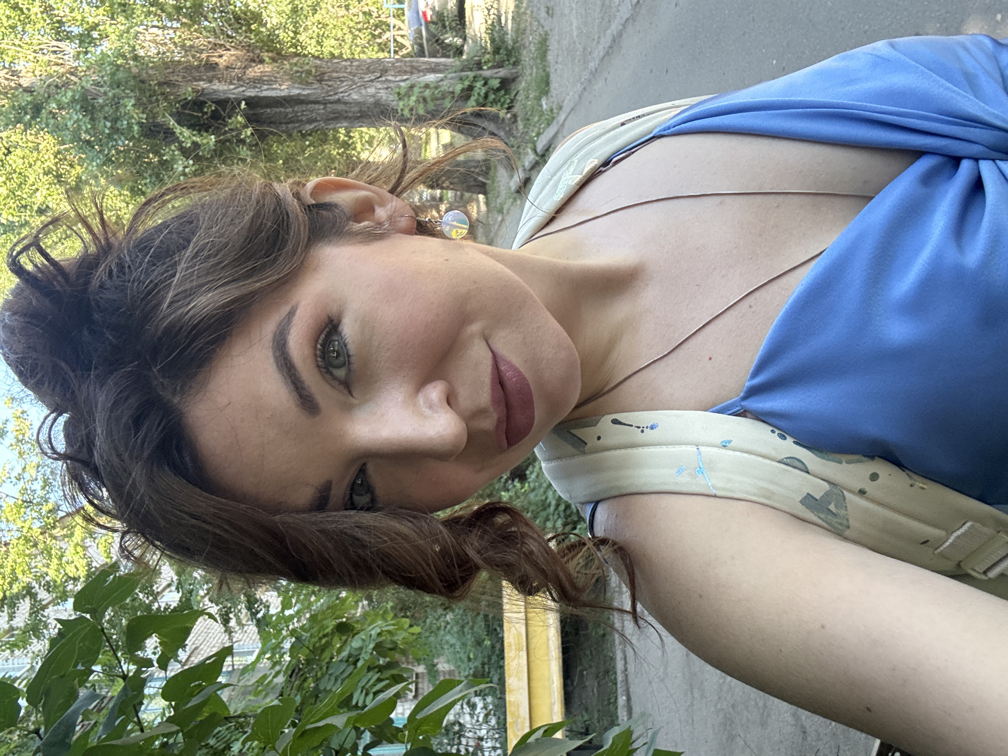 A woman with dark curly hair tied up in a messy bun, with loose curls framing her face, is taking a selfie outdoors. She is wearing a blue dress with a low neckline and a light-colored backpack with a paint-splattered design. She has on a delicate necklace and a dangling earring. She is looking at the camera with a slight smile. The background shows trees, a building, and a yellow bench, suggesting an urban park or residential area in Kryvyi Rih, Ukraine. The lighting indicates it is daytime, likely during the warmer months.