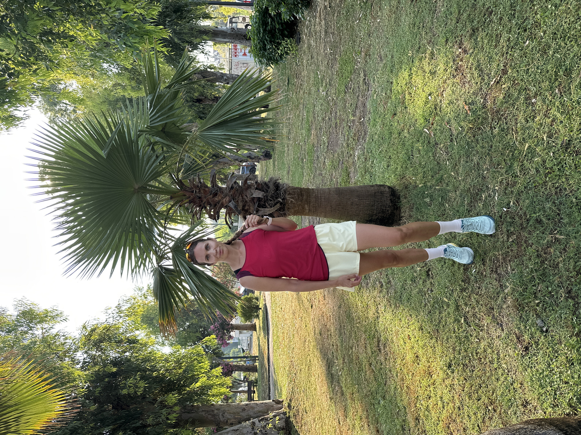 A full-body, medium-shot photograph features a woman standing in a grassy park area, likely in Fethiye, Turkey. She is positioned slightly to the right of center, in front of a palm tree. The woman has light skin, brown hair braided down her back, and is wearing sunglasses pushed up on her forehead. Her attire consists of a dark pink tank top and light yellow shorts, white socks, and light blue athletic shoes. Her left hand is holding the braid of her hair, and she is looking directly at the camera with a neutral expression.
The environment appears to be a park with a large palm tree dominating the center-right of the frame. Behind the woman, tall trees with green foliage line the background, with sunlight filtering through them. The ground is covered in grass, with some patches of bare earth and fallen leaves. In the further background, out of focus, are park benches, some structures that might be part of a playground or public facilities, and possibly vehicles. The overall impression is an outdoor setting in a populated area, possibly a municipal park. The lighting suggests it is late afternoon or early morning, with the sun casting long shadows.