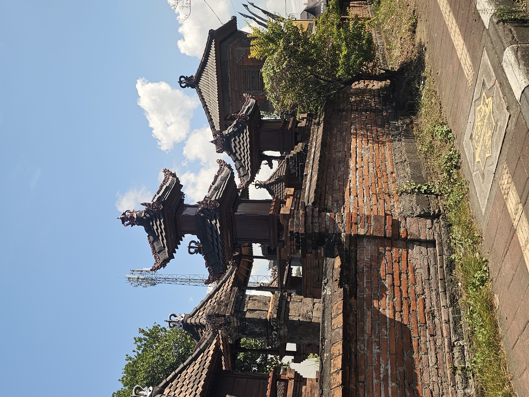 The image displays a traditional Balinese architectural style in the city of Sukawati, Indonesia. In the foreground, a brick wall with a rough texture runs horizontally across the frame, adorned with decorative stonework and multiple tiered roofs. Several multi-story structures with intricate carvings and pagoda-like roofs are visible behind the wall. A green tree stands to the right, casting a shadow on the paved ground. In the background, a cell tower stands tall against a clear blue sky dotted with white clouds. The scene is bathed in bright sunlight, indicating a daytime setting. The overall impression is one of cultural heritage and a blend of traditional and modern elements in the urban landscape of Sukawati.