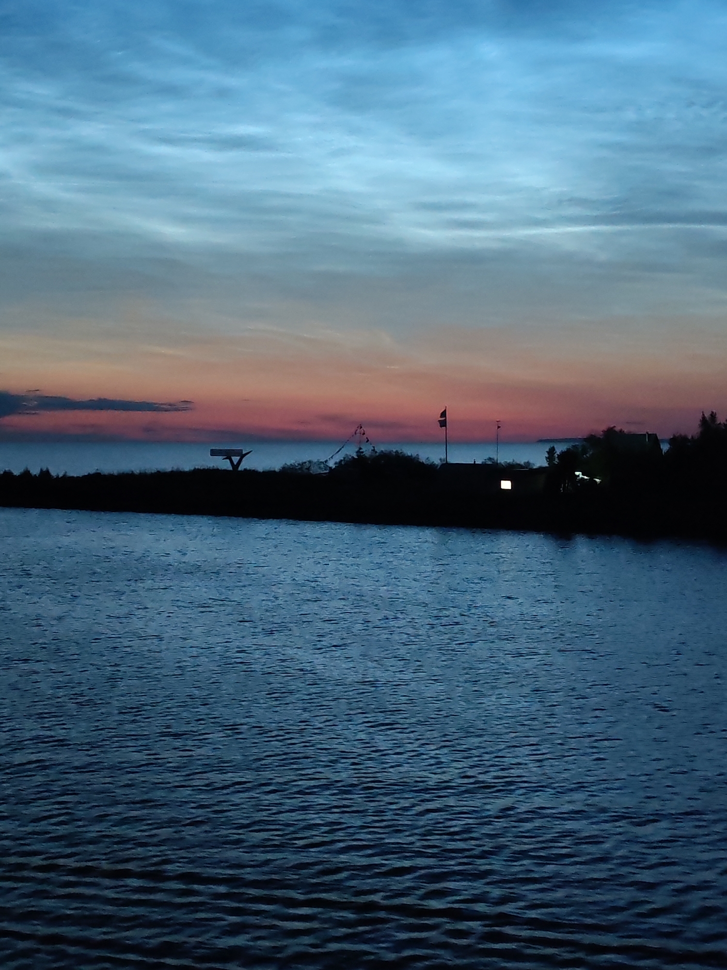 The image captures a twilight scene over a body of water, likely a lake or bay, in Ozernoe Ust'e, Russia. The sky is a gradient of deep blue at the top, transitioning to lighter blues and then a band of soft pink and orange near the horizon, indicating sunset or sunrise. Wispy clouds streak across the sky. In the middle ground, a dark, silhouetted shoreline is visible, dotted with trees and structures. A prominent object on the shore is a distinctive, abstract sculpture or landmark with a triangular base and a flat, horizontal top. Further to the right, a building with a lit window is silhouetted, and two flags on poles are visible, one darker and larger than the other. The foreground is dominated by the dark, rippling surface of the water, reflecting the dim light of the sky. No people are visible in the image. The overall atmosphere is serene and tranquil.