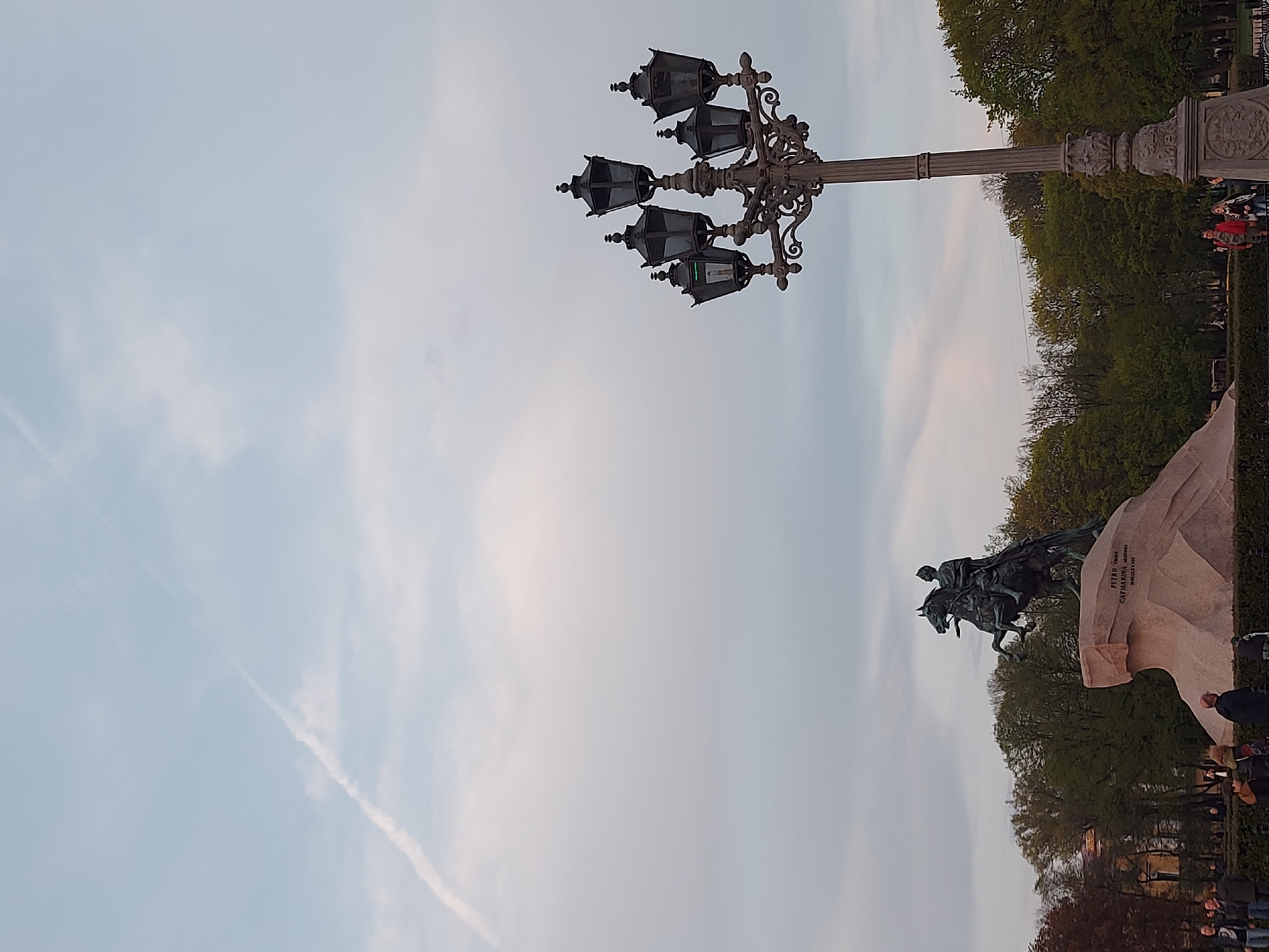 The image shows the Bronze Horseman, an equestrian statue of Peter the Great in Senate Square, Saint Petersburg, Russia. A lamppost is visible in the upper center of the image, and trees and people surround the statue. The sky is cloudy and light blue.
