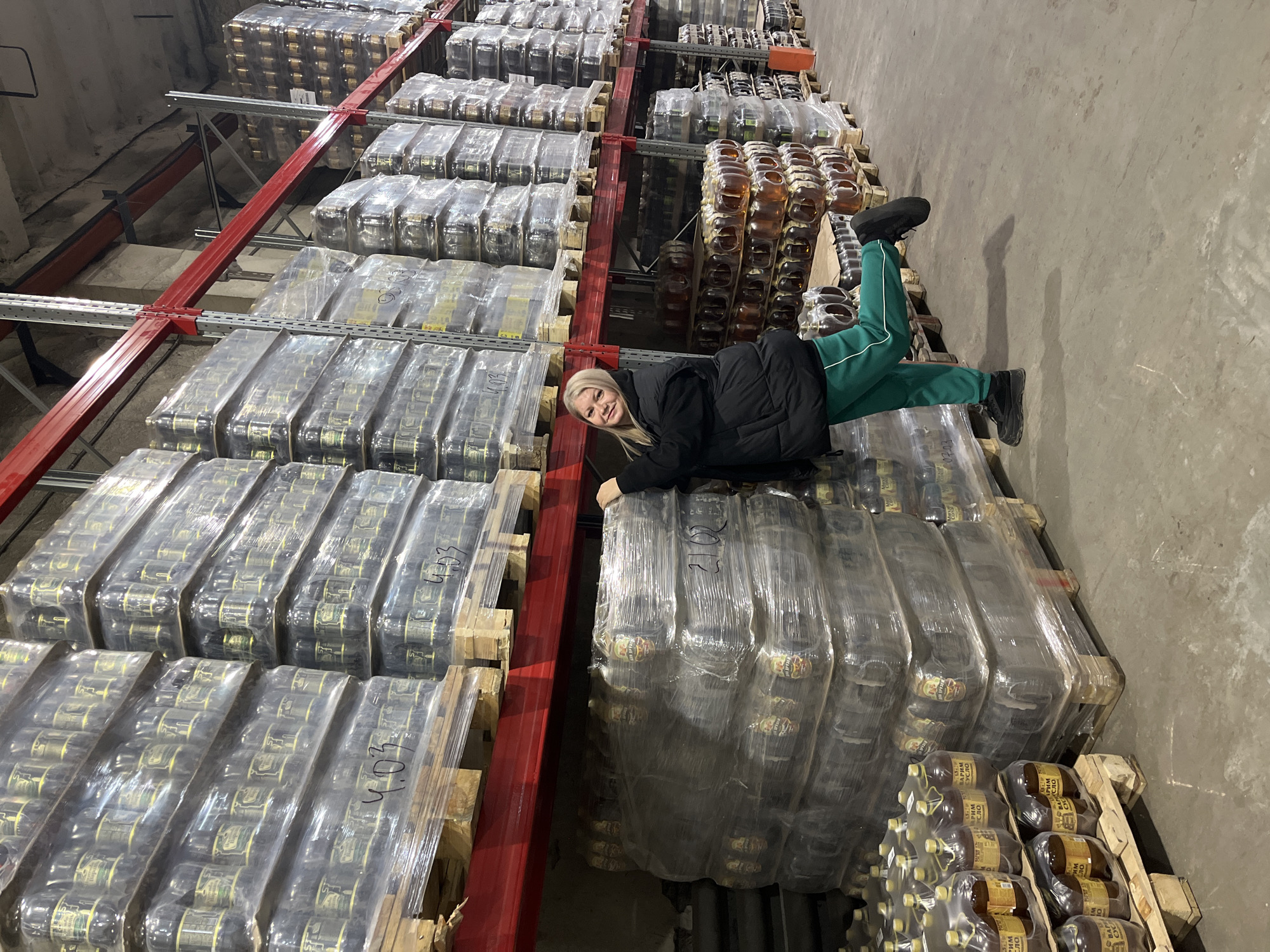 A female subject is depicted within an industrial warehouse, likely located in or serving the Izhevsk region of Russia, identifiable by the presence of "ВАРЯМ СУСЛО" (Varyam Suslo) branded bottled beverages, a product of the Udmurt Republic. The warehouse features multiple rows of grey metal shelving units with red horizontal support beams, extending into the background and upwards. These shelves are densely packed with wooden pallets holding stacks of plastic-wrapped cases containing numerous plastic bottles with brownish liquid. Handwritten numerical markings, such as "4.03" and "21.03," are visible on some of the plastic wraps.
The subject, a blonde-haired female appearing to be a young adult, is positioned in the foreground on the left. She wears a black puffer jacket, green track pants with a white stripe, and black athletic shoes. She is smiling towards the camera, leaning her torso against a stack of packaged goods, with her right hand resting on the topmost case. Her left leg is bent at the knee and raised off the ground. The floor is a grey concrete slab, and the upper section of the warehouse reveals concrete walls, ceiling, and metal structural components.