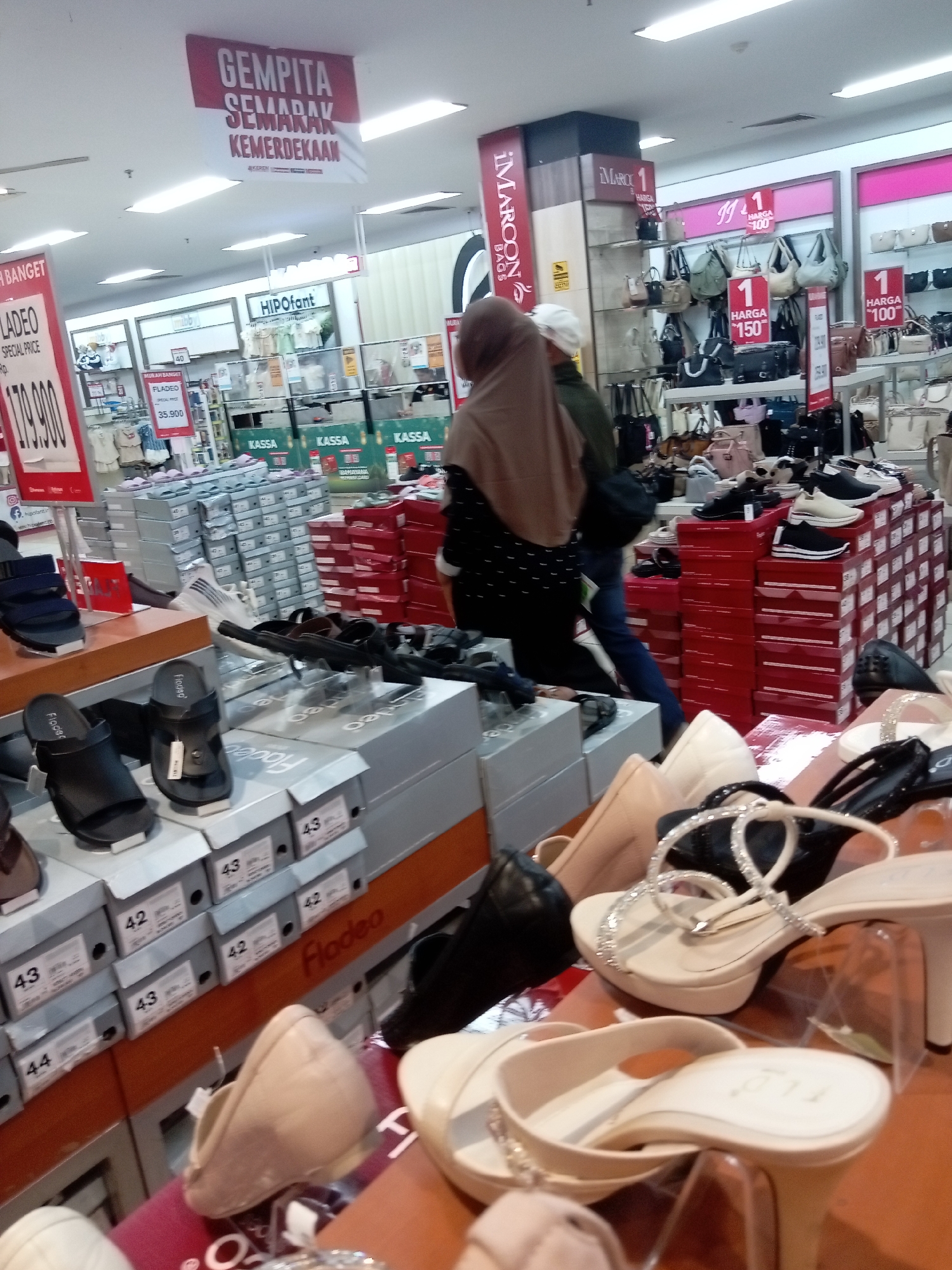 This image depicts the interior of a shoe store, likely in Batam, Indonesia, given the presence of Indonesian text on signage. The store appears to be a department store or a large retail outlet specializing in footwear and bags.
In the foreground, a variety of women's shoes are displayed on shelves and tables. These include high heels, sandals, and flats, with some shoes presented in their original boxes. The shoe boxes are stacked neatly, with labels indicating sizes and brands. Brands like "Fladeo" are visible.
In the mid-ground, a display of handbags is prominent, with many bags hanging on shelves. Several bags are displayed with price tags indicating "HARGA 100" (Price 100) and "HARGA 150" (Price 150), suggesting these are currency denominations, likely Indonesian Rupiah. The brand "iMAROON BAGS" is also visible. In the background, more merchandise is visible, including clothing items on racks.
Two individuals are partially visible in the scene. One person, wearing a beige hijab and a patterned top, is looking towards the shoe displays. Another person, wearing a white cap and a dark jacket, is also in the vicinity, possibly browsing.
The overall environment is brightly lit by fluorescent ceiling lights, typical of a retail setting. Signs are present throughout the store, with a prominent red sign at the top reading "GEMPITA SEMARAK KEMERDEKAAN" (Festive Independence) in Indonesian, indicating a promotional event tied to Indonesian Independence Day. Other signs in Indonesian like "HIPOfant" and "KASSA" (Cashier) are also visible. The store appears to be well-stocked and organized, with a focus on presenting merchandise attractively to customers.