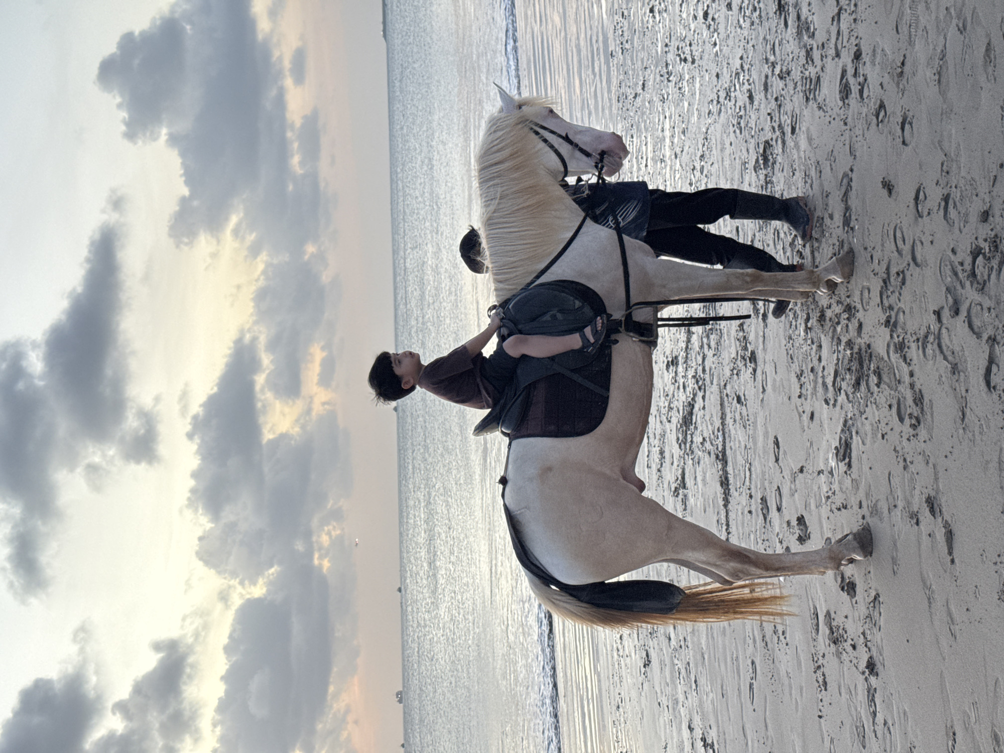 A daytime, medium, full shot shows a pale horse with a child riding it on a sandy beach in Kuta Selatan, Indonesia. The horse faces right. A handler stands behind the horse with black boots and pants. The child wears a brown shirt and dark shorts and sandals. The beach is covered in footprints. The ocean is in the background with a sky with clouds.