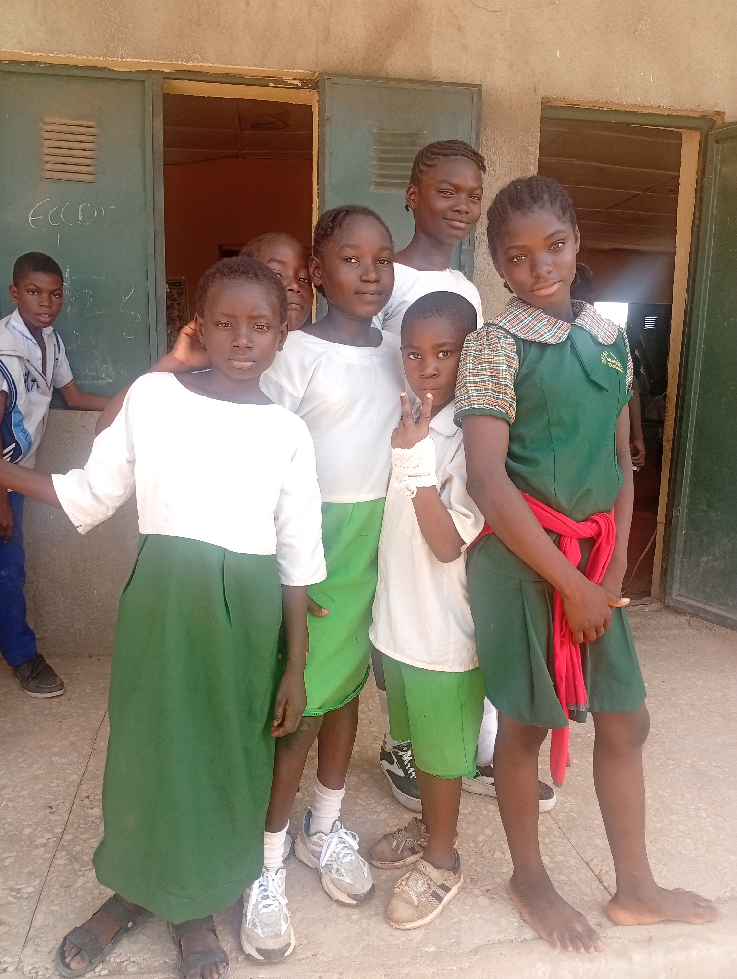 A group of seven children, primarily girls, stand outdoors in front of a light-colored building with two open, green-painted metal doors, likely a school in Narayi, Nigeria. The children are posing for a photograph.
In the foreground, from left to right, a young girl wears a white long-sleeved top and a dark green skirt, standing barefoot with a direct gaze at the camera. Next to her, a boy in a white short-sleeved shirt and green shorts makes a peace sign with his right hand, which is wrapped in a white bandage. To his right, a girl wears a dark green dress with a plaid collar and sleeves, cinched at the waist with a red fabric sash or scarf. She is also barefoot, and a badge on her dress reads "GREAT MERIT SCHOOL".
Behind these children, a girl in a white top and green shorts is partially visible near the center. To her right, a taller girl in a white top looks slightly off-camera. From the left open doorway, a boy in a white and blue outfit peeks out into the frame. The children display a mix of footwear, with some wearing sneakers or sandals, while others are barefoot.
The ground is a light, paved surface. The green metal doors behind them have visible chalk-like markings, including "FCCC" and numbers "123". The bright and even lighting suggests it is daytime, likely a sunny day. The children's expressions are mostly neutral or subtly smiling.