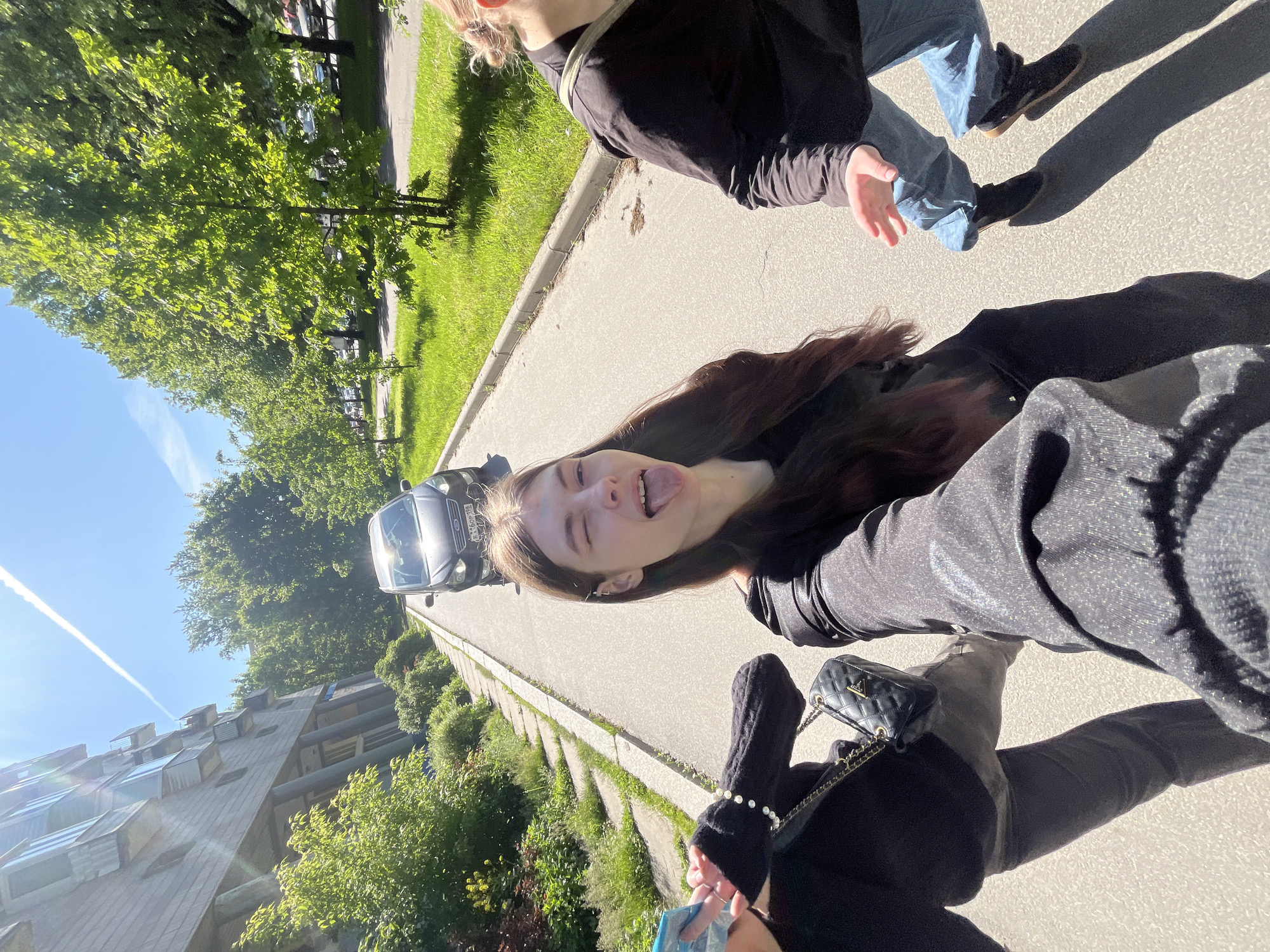 This is an outdoor selfie-style photograph taken on a bright, sunny day, captured from a slightly high angle.
Two young women are the main subjects. In the foreground, one woman is taking the photo, partially obscuring her hand holding the camera. She has long, dark hair, is wearing a dark top, a pearl bracelet on her left wrist, and a small black quilted handbag with a visible "GUESS" logo. She is making a playful expression, sticking out her tongue. Behind her, another woman is partially visible, wearing a black top and light-colored jeans.
They are positioned on a paved surface, likely a sidewalk or road, with a grassy verge and lush green trees lining one side. In the mid-ground, a dark-colored car, possibly a Ford, is parked. Part of its Russian license plate, "М46P", is discernible, suggesting a region code like '78' or '178' (Saint Petersburg). Beyond the car and trees, residential buildings are visible under a clear, bright blue sky with a prominent contrail.
The time of day appears to be late morning or early afternoon, indicated by the strong sunlight and distinct shadows cast on the ground. The overall mood is casual and cheerful, conveyed by the foreground subject's playful expression.