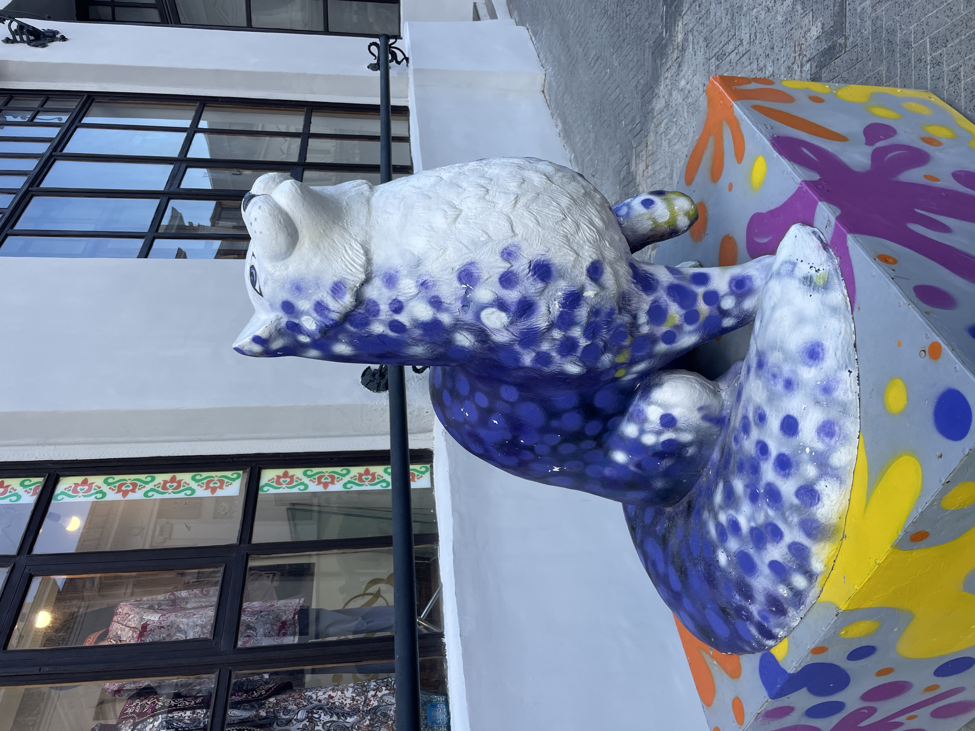 The image showcases a vibrant, stylized animal sculpture positioned outdoors against a modern building backdrop. The main subject is a large sculpture, resembling a lounging cat or similar creature, painted predominantly white with numerous blue and purple polka dots. The sculpture's head is turned to the left, and its body extends horizontally, resting on a multi-faceted base.
This base is a grey, geometric block heavily spray-painted with splashes and spots of yellow, orange, purple, and blue, giving it a lively, graffiti-like appearance.
In the background, a white building with large, dark-framed windows rises. Reflections in the windows reveal glimpses of the surroundings. Above some lower windows, a distinctive decorative border is present, featuring repeating patterns of green and red, possibly depicting heart-like or floral motifs. Through the windows, various patterned textiles or fabrics are visible, suggesting the building houses a shop. A slender black metal pole stands vertically to the left of the sculpture. To the far right, a textured grey brick wall is partially visible.
The scene is set during daylight hours, indicated by the bright and clear lighting. There is no specific activity taking place, and no people are visible. No text is discernible within the image. The decorative elements and urban setting align with the provided context of Kazan, Russia, suggesting an outdoor public art display.