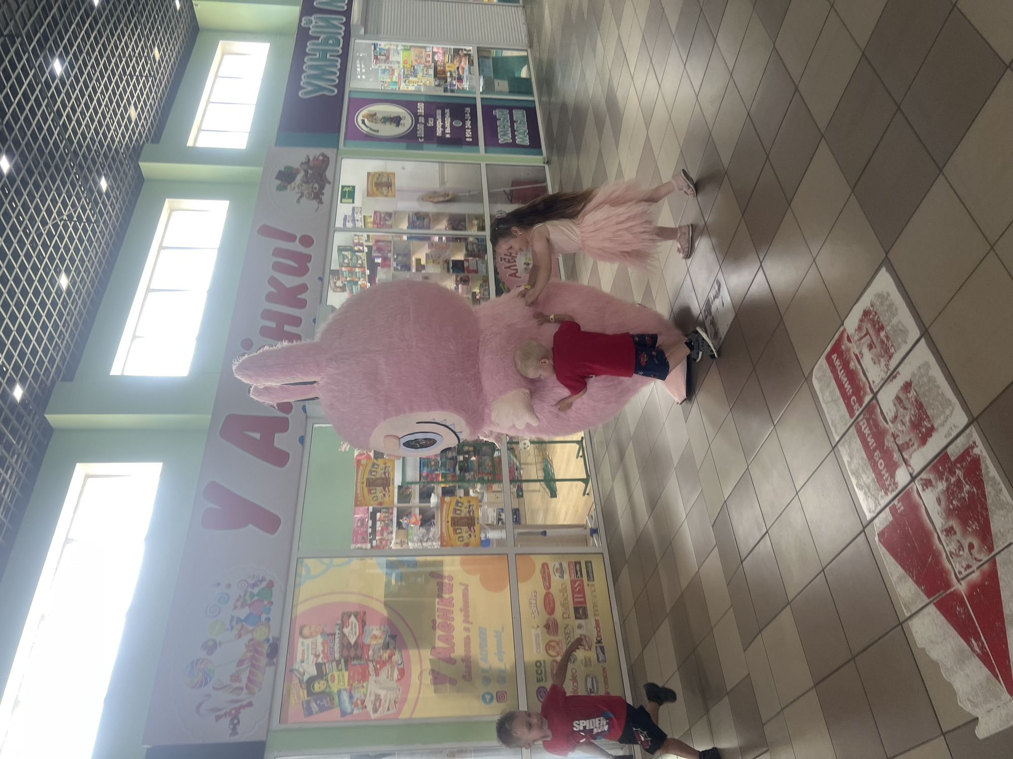 In an indoor shopping mall with a tiled floor and a grid-like ceiling with fluorescent lighting, a large pink bunny mascot is interacting with three young children. To the left, a young boy in a red t-shirt with a Spider-Man logo and black shorts is running, looking back towards the mascot. In the center, a younger boy wearing a red t-shirt and patterned shorts is standing next to the mascot, appearing to be touching its leg. To the right, a young girl in a pink tutu dress and sandals is reaching out to hug or touch the mascot. The mascot is a tall, fluffy pink costume with large ears and a friendly expression. Behind the mascot are storefronts, one with a large sign featuring colorful candies and the text "У Алёнки!", and another with the text "УМНЫЙ М". Several children's toys and products are visible inside the storefronts. A red and white arrow graphic is on the floor in the foreground, pointing towards the right side of the image.