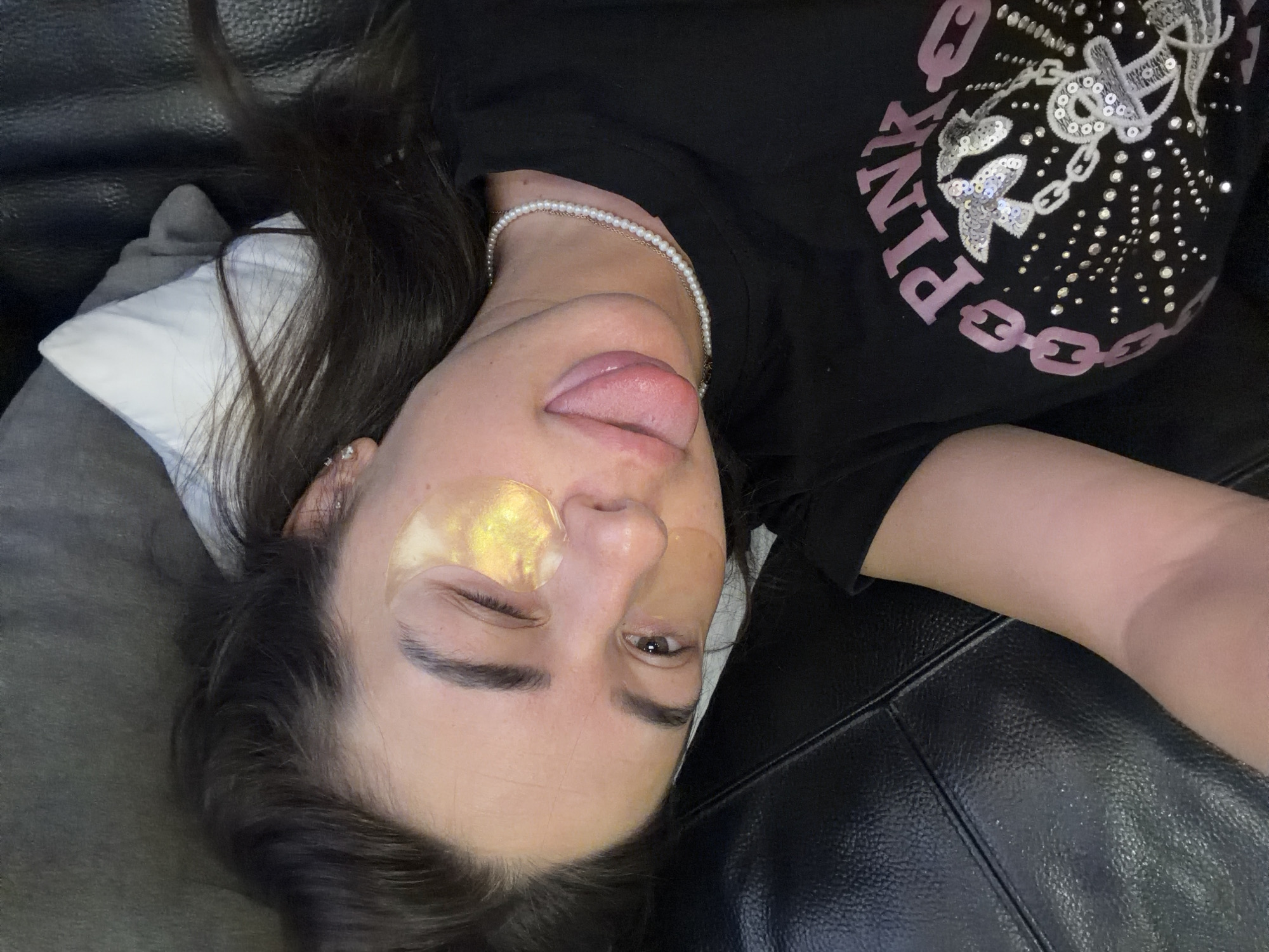 A young adult female, with dark hair, is lying on a black leather sofa. She is wearing a black t-shirt with pink lettering and sequins, and a pearl necklace. She has gold under-eye patches on, and her left eye is closed. Her tongue is sticking out, and she is looking towards the camera. The environment appears to be an indoor setting, possibly a living room in Novi Iskar, Bulgaria.