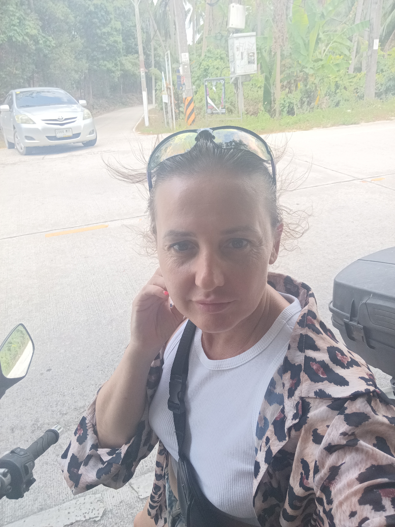 A middle-aged woman is the central subject of a selfie taken outdoors. She is wearing a white ribbed tank top and a tan and black leopard-print jacket. Sunglasses rest on top of her head. The woman's right hand is raised near her face.
She appears to be seated on a motorcycle, evident from the mirror and handlebar visible on the left. To the right, a large black box, possibly attached to the motorcycle, can be seen. The backdrop features a paved road with yellow road markings. A silver car is parked on the left side of the road. Green vegetation and trees line the roadside. Also present is an electrical box on a pole. The photo's location is in Thong Sala, Thailand.