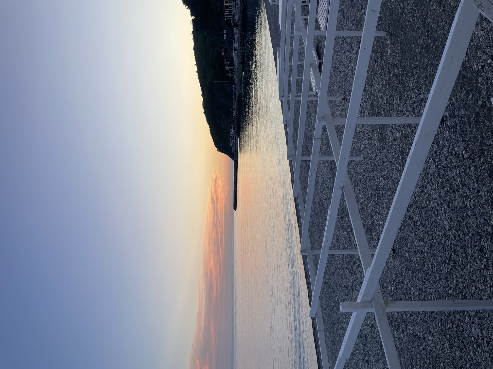 The image presents a landscape taken at what appears to be the coast of Olginka, Russia, during either sunrise or sunset. A body of water, likely the sea, reflects the colors of the sky, transitioning from blue to orange. A strip of land, possibly a hillside covered in vegetation, is visible on the left side. A structure built of white beams and poles, possibly part of a shelter, is present on the right side, extending into the scene. A stony ground is visible along the right side of the frame.