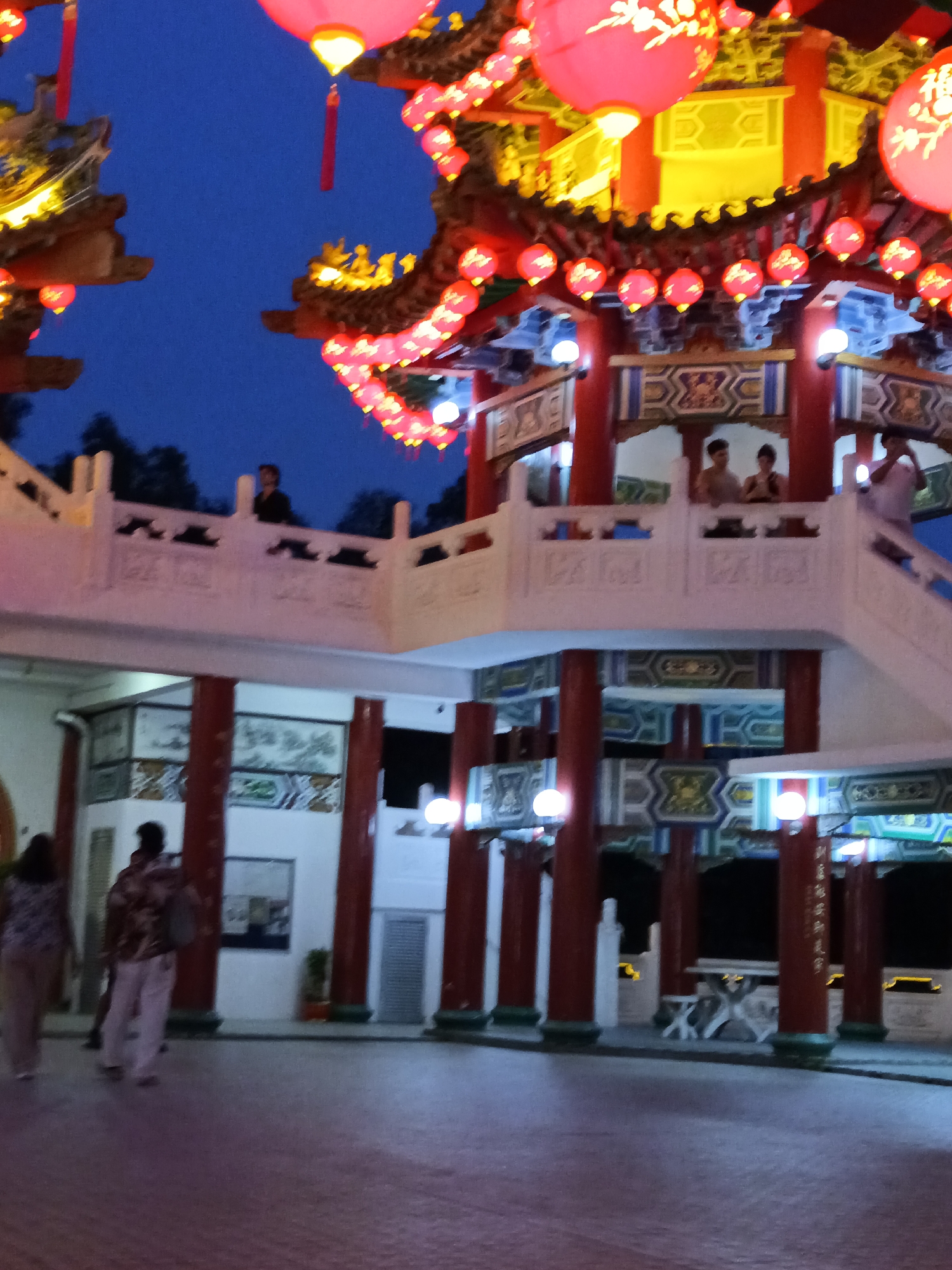 The image depicts the Thean Hou Temple in Kuala Lumpur, Malaysia, at dusk. The temple's architecture is characterized by red columns, ornate roofs, and numerous red lanterns, some of which are illuminated. A multi-tiered structure with walkways and staircases is prominent, with people visible on different levels. In the foreground, two individuals are walking away from the camera on a paved surface. The sky is a deep blue, indicating the transition from daylight to night. The scene is illuminated by artificial lighting, including the red lanterns and lights within the temple structure.