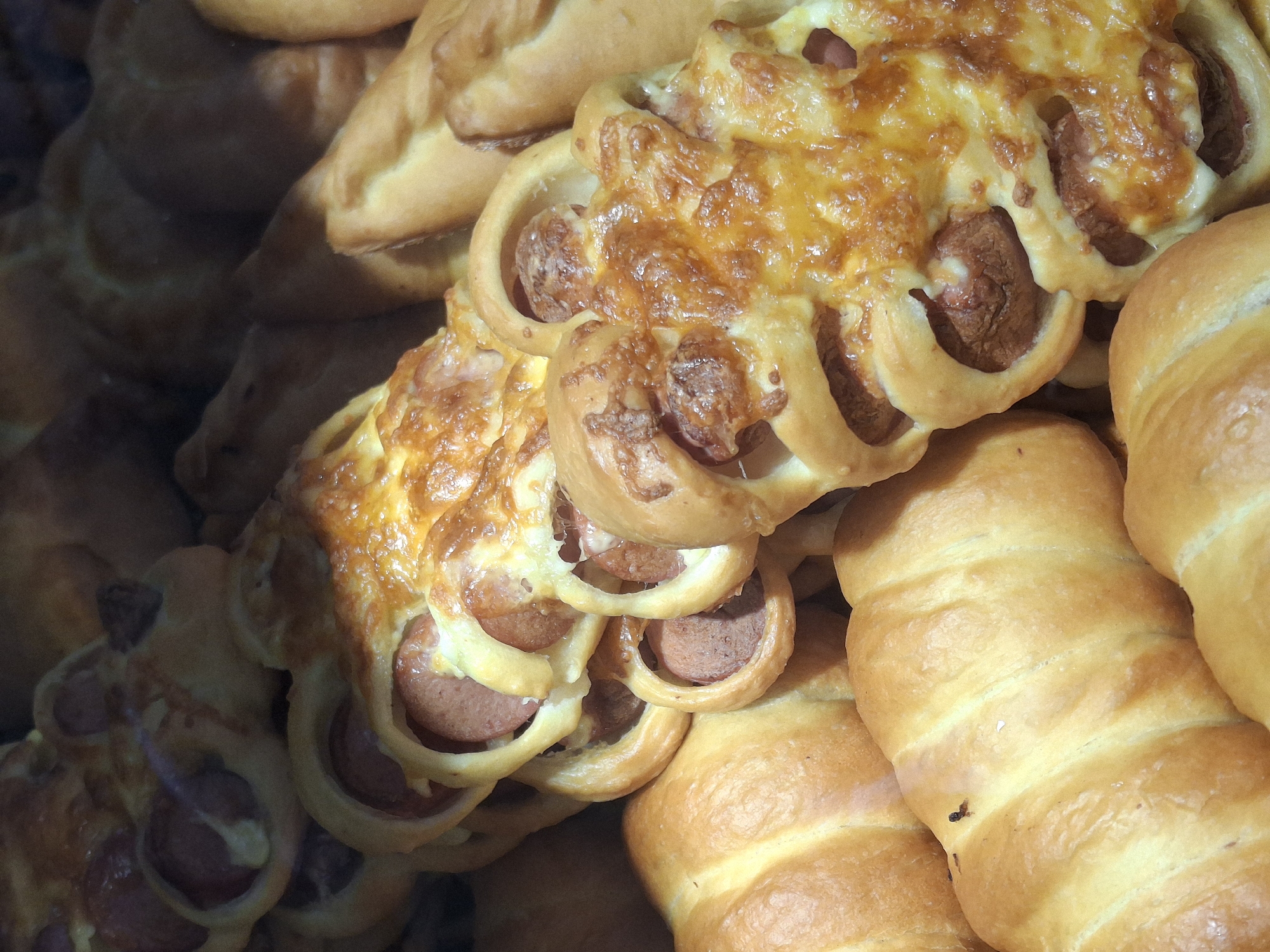A close-up image shows a stack of baked goods, likely in a bakery or food display case. Prominently featured are pastries with sausage slices embedded in a lattice of dough, topped with melted cheese. Adjacent to these are several rolled pastries, possibly filled with meat or cheese. The scene depicts food items ready for sale or consumption. The location is assumed to be in Chaikovsky, Russia.
