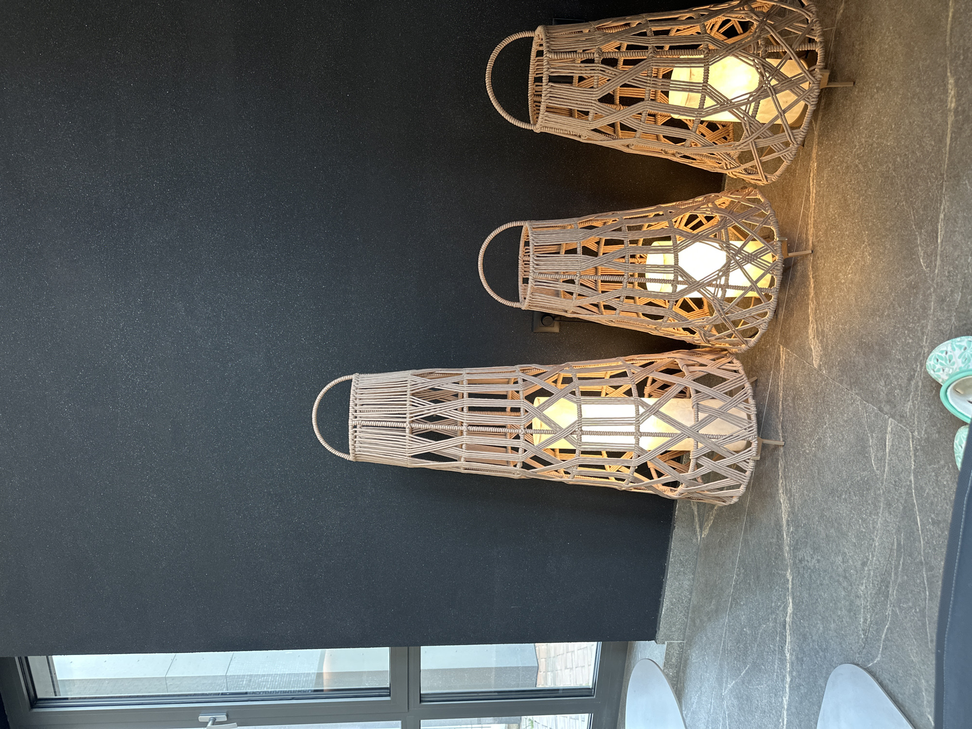 The image shows three cylindrical lamps made of woven rope hanging on a dark wall. Each lamp has a handle and a light bulb inside, which illuminates the lamp and surrounding area. The wall is smooth and dark in color. Below the wall, there is a window with a gray frame. To the right of the wall, there is a gray tiled floor. Part of the bottom of two chairs can be seen. The location is Kazanlak, Bulgaria.