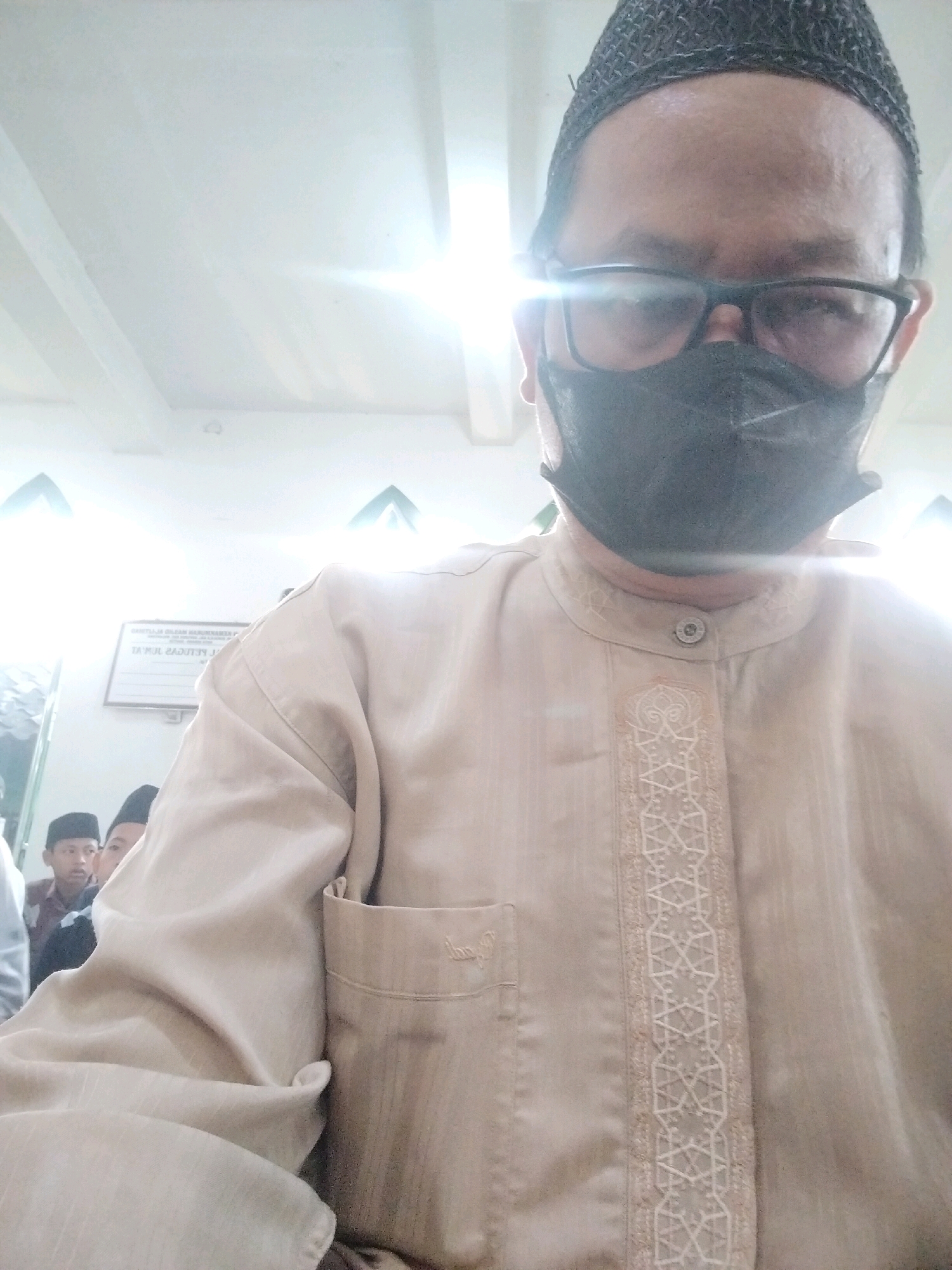 A medium shot captures a man wearing a black skullcap, eyeglasses, and a black face mask. He is looking directly at the camera, with his head tilted slightly to the right. He is wearing a light brown tunic with a white embroidered pattern down the front. In the background, two young boys wearing skullcaps are visible, and a sign with Indonesian text is displayed on the wall. The environment appears to be indoors, possibly a mosque or a community center in the city of Serang, Indonesia, suggested by the presence of the sign and the attire of the individuals. The lighting in the room is bright, with some reflections visible on the ceiling.