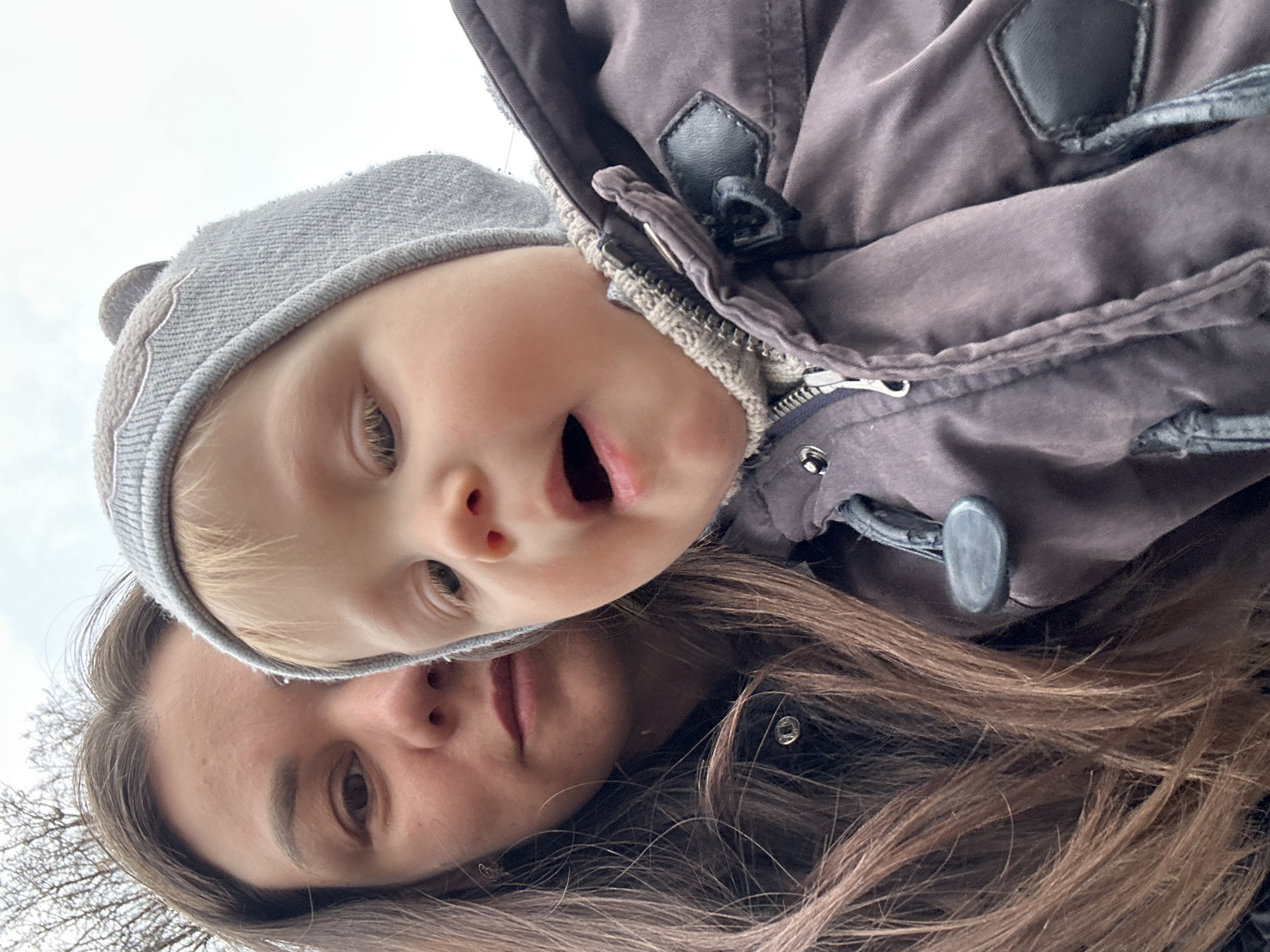 A close-up, angled-upward shot depicts a woman and a young child, possibly in the city of Chaikovsky, Russia (based on assumption given location information). The child, wearing a gray knit hat, is positioned above the woman, with his head tilted. The child's mouth is open. The woman's face is visible beneath the child. Her long brown hair frames her face. She appears to be wearing a dark jacket. Bare trees are visible in the background.