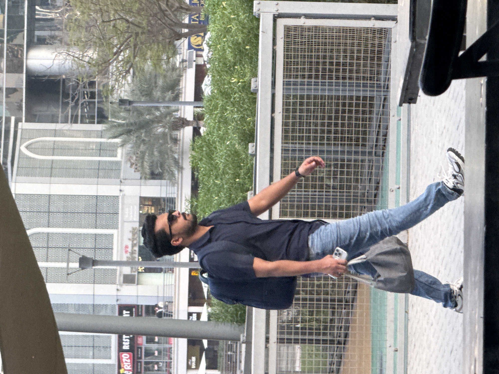 A medium shot captures a man walking outdoors in Dubai, United Arab Emirates. The man, wearing sunglasses, a dark short-sleeved polo shirt, blue jeans, and athletic shoes, carries a dark backpack and a grey messenger bag. He holds a smartphone in his right hand. In the background, a modern building with arched windows and several palm trees are visible. The man is walking along a paved pathway beside a metal mesh fence.