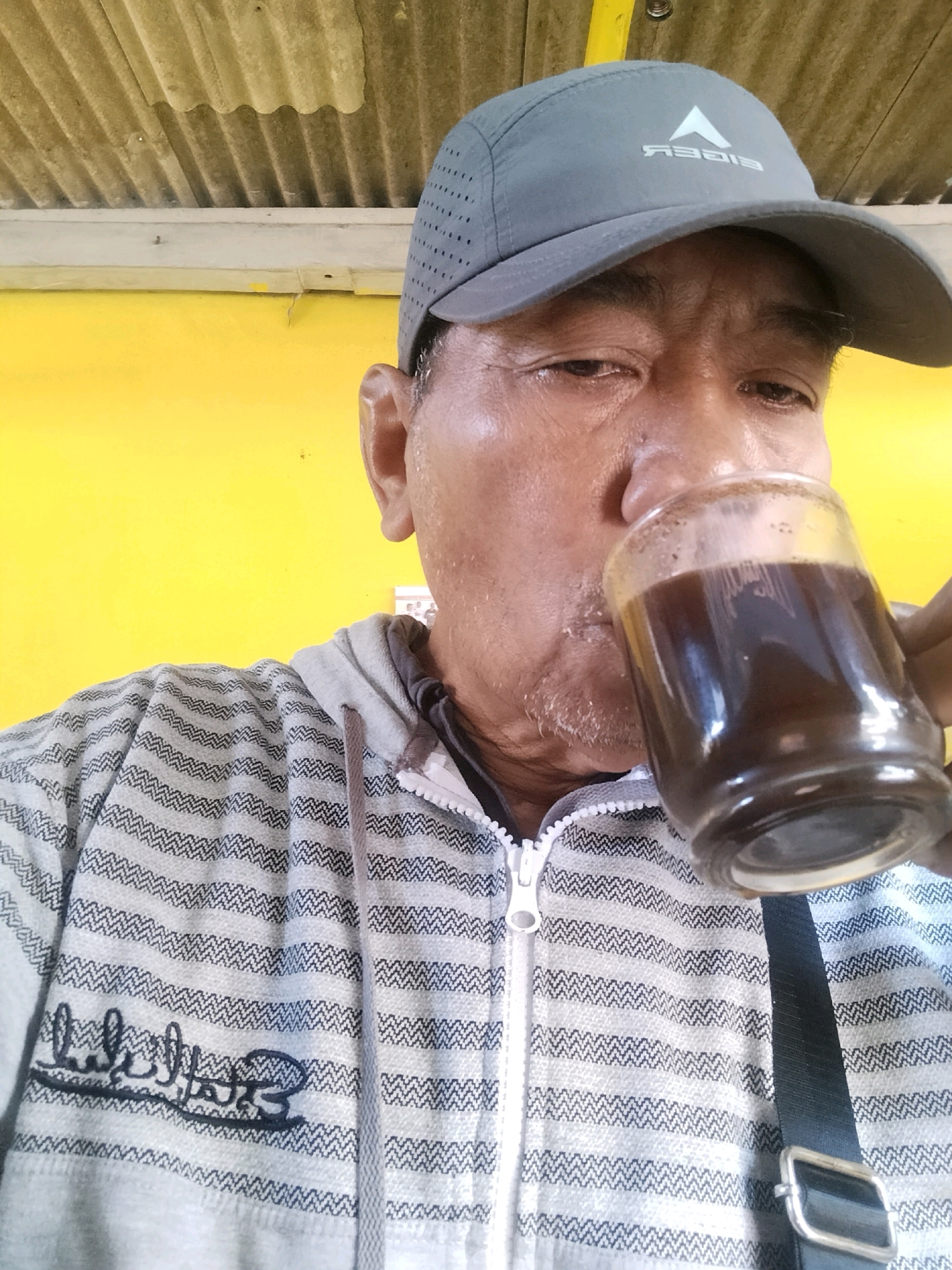 A close-up, waist-up, indoor shot shows an elderly East Asian man wearing a gray baseball cap and a gray and black striped zip-up hoodie. The man is holding a clear glass with dark liquid, presumably coffee, to his lips, and appears to be drinking from it. His head is tilted to the right, and his eyes are partially closed. The background is a bright yellow wall, and above that, a corrugated metal ceiling. A black strap from a bag is visible diagonally across the man's chest. The lighting is natural and appears to be from above. The overall impression is that the man is enjoying a beverage in a casual, outdoor setting.