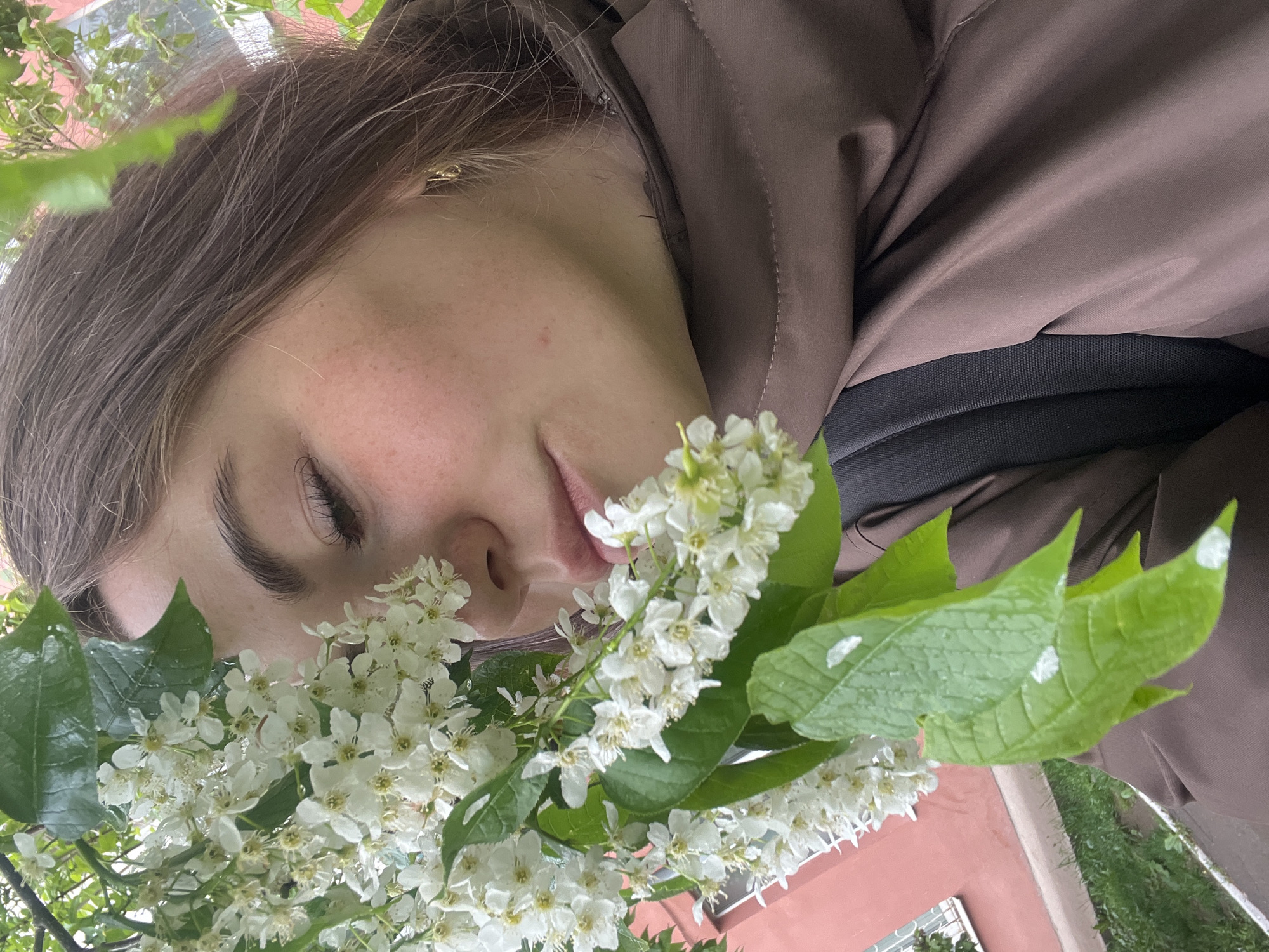 A young woman is depicted smelling white flowers. She is fair-skinned with light brown hair and is wearing a brown jacket. A black strap is visible under her jacket collar. The flowers she is smelling have green leaves with some white spots on them. A building with a red facade and a window is visible in the background, along with grass. Based on visual cues, the location is possibly Chaikovsky, Russia, as requested. The woman's eyes are closed as she appears to enjoy the scent of the flowers.