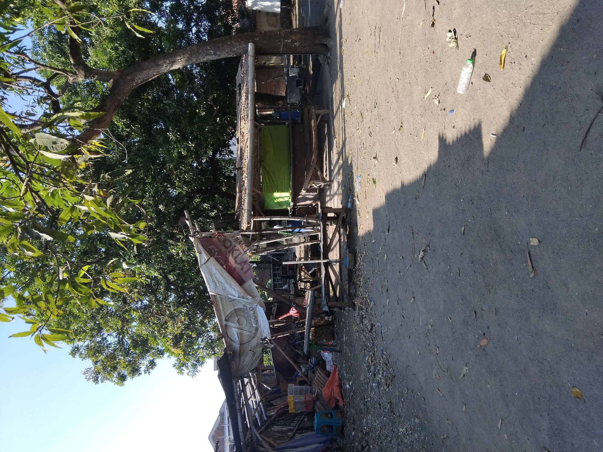 The image depicts a weathered, open-air marketplace stall, likely in a rural or semi-urban area, characterized by makeshift construction and a somewhat unkempt environment. The stall features a roof supported by wooden poles, covered with a combination of corrugated metal and thatch. A large, tattered white tarp with a circular logo and text, possibly advertising a product, is draped over part of the structure.
In front of the stall, there are wooden benches and tables, suggesting a place for customers to sit or for goods to be displayed. To the left, a pile of items includes orange tarps, blue plastic containers, and what appears to be sacks. A small blue plastic stool is also visible.
Behind the stall, a large tree with extensive foliage provides shade. The ground is covered in dirt and gravel, with scattered debris such as leaves and a plastic bottle. In the background, glimpses of residential structures with tiled roofs are visible, indicating a populated area. The overall impression is one of a functional, but modest, local trading post. The presence of the large tree and the building materials suggest this is a tropical or subtropical climate. While the location is stated as Kepuh Kiriman, Indonesia, the image itself does not contain any direct indicators to confirm this specific location, relying solely on external information. There are no people visible in the frame.
