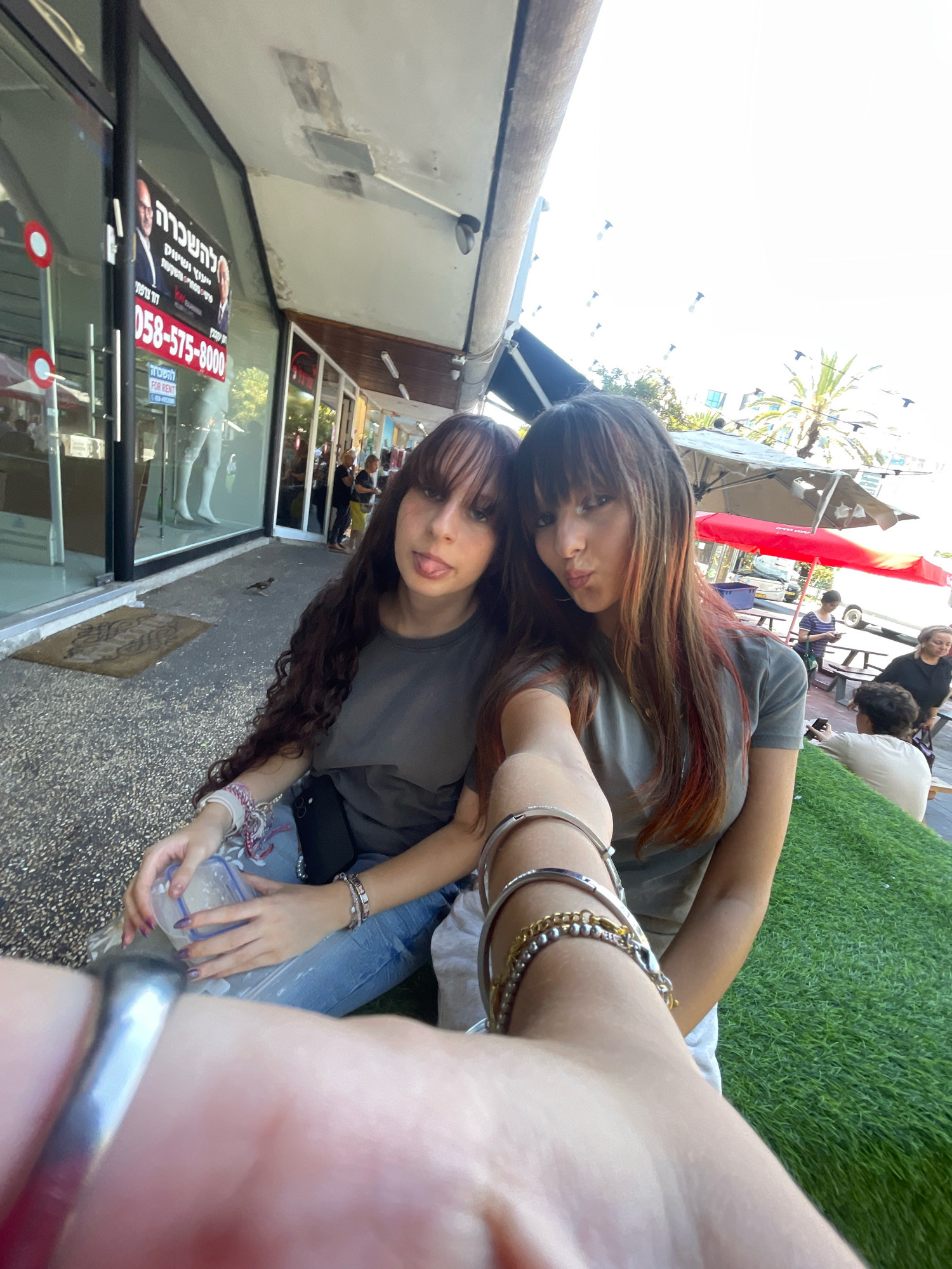 Two young women are taking a selfie outdoors in front of a storefront. The woman on the left has long, dark, curly hair and is sticking her tongue out. She is wearing a grey t-shirt and blue jeans and is holding a clear plastic container. The woman on the right has long, dark hair with streaks of red and is puckering her lips. She is wearing a grey t-shirt. Both women are wearing bracelets. In the background, there are tables and chairs, an umbrella, and other people. A sign on the storefront displays text in Hebrew, including a phone number, and images of two men. The ground is a speckled asphalt surface. The location is Raanana, Israel.