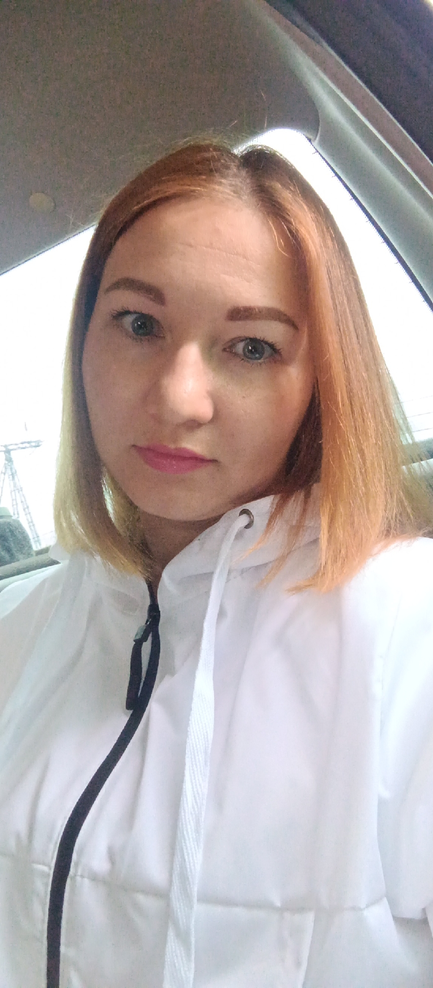 A woman with auburn hair and blue eyes is shown in a close-up selfie within the interior of a vehicle. She is wearing a white jacket with a black zipper and white drawstrings. The backdrop suggests a city environment, with an out-of-focus structure possibly being a utility tower visible through the car window.