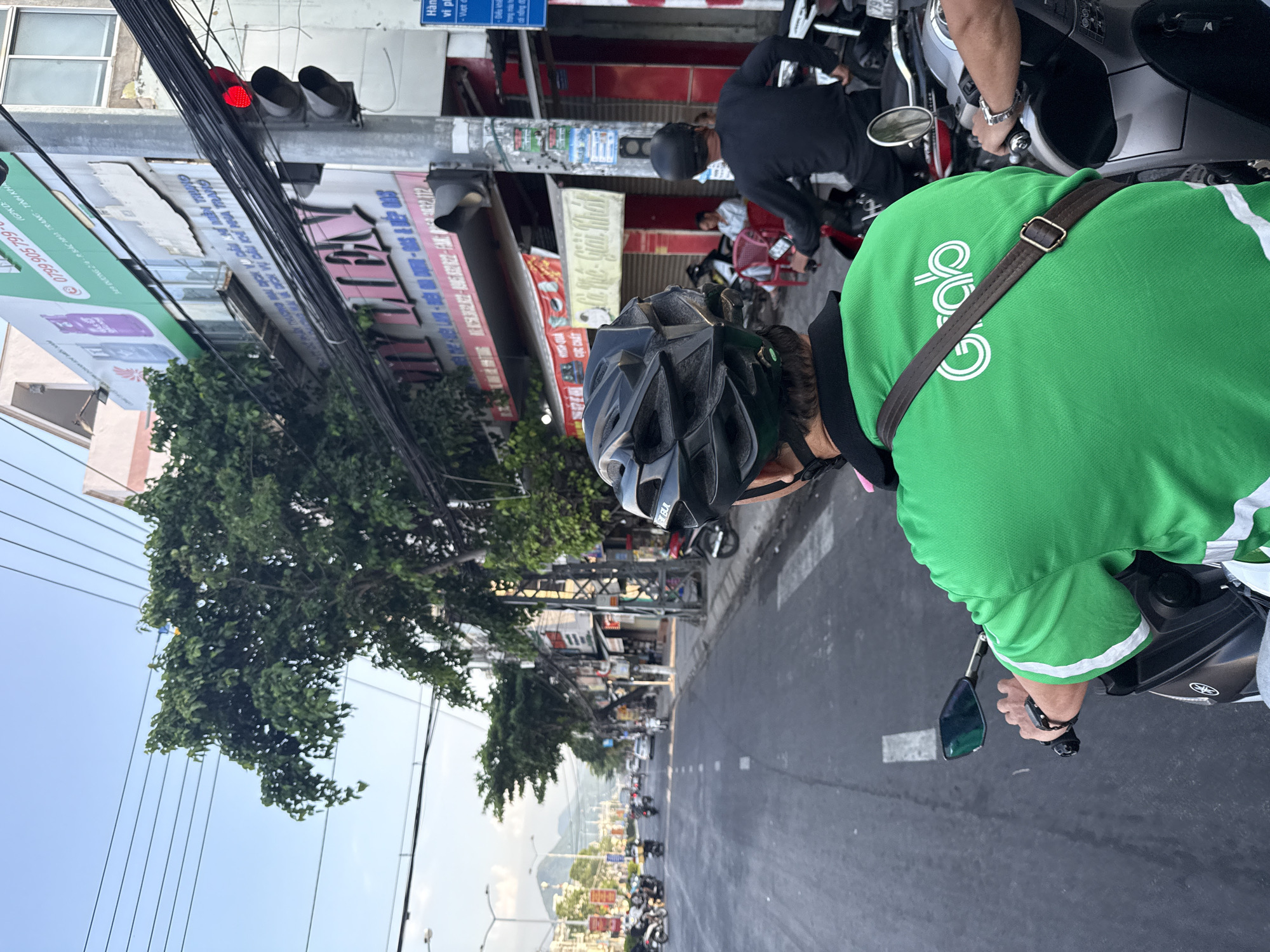 The image depicts a street scene in Bac Nha Trang Ward, Vietnam. In the foreground, a person wearing a bright green shirt with the "Grab" logo and a black helmet is riding a motorcycle. The rider's left hand is visible on the handlebars. To the right, another person, also wearing a black helmet, is riding a motorcycle. In the background, there are buildings with signage, trees, overhead power lines, and other vehicles on the road, including motorcycles and a car. The sky is clear with some clouds, and the scene appears to be during daylight hours. A traffic light with a red light illuminated is visible on a pole.