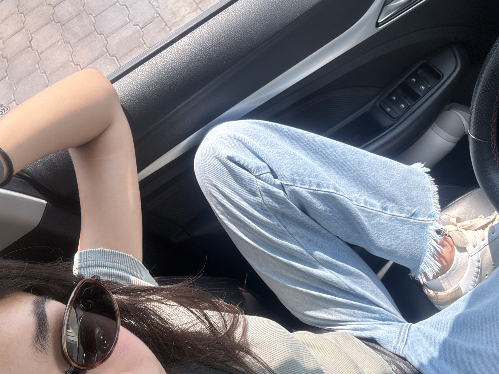 A selfie taken from inside a car. The subject is wearing sunglasses and light blue jeans. The car's interior features black plastic trim and controls. A white sneaker is visible near the steering wheel. The location is unspecified.