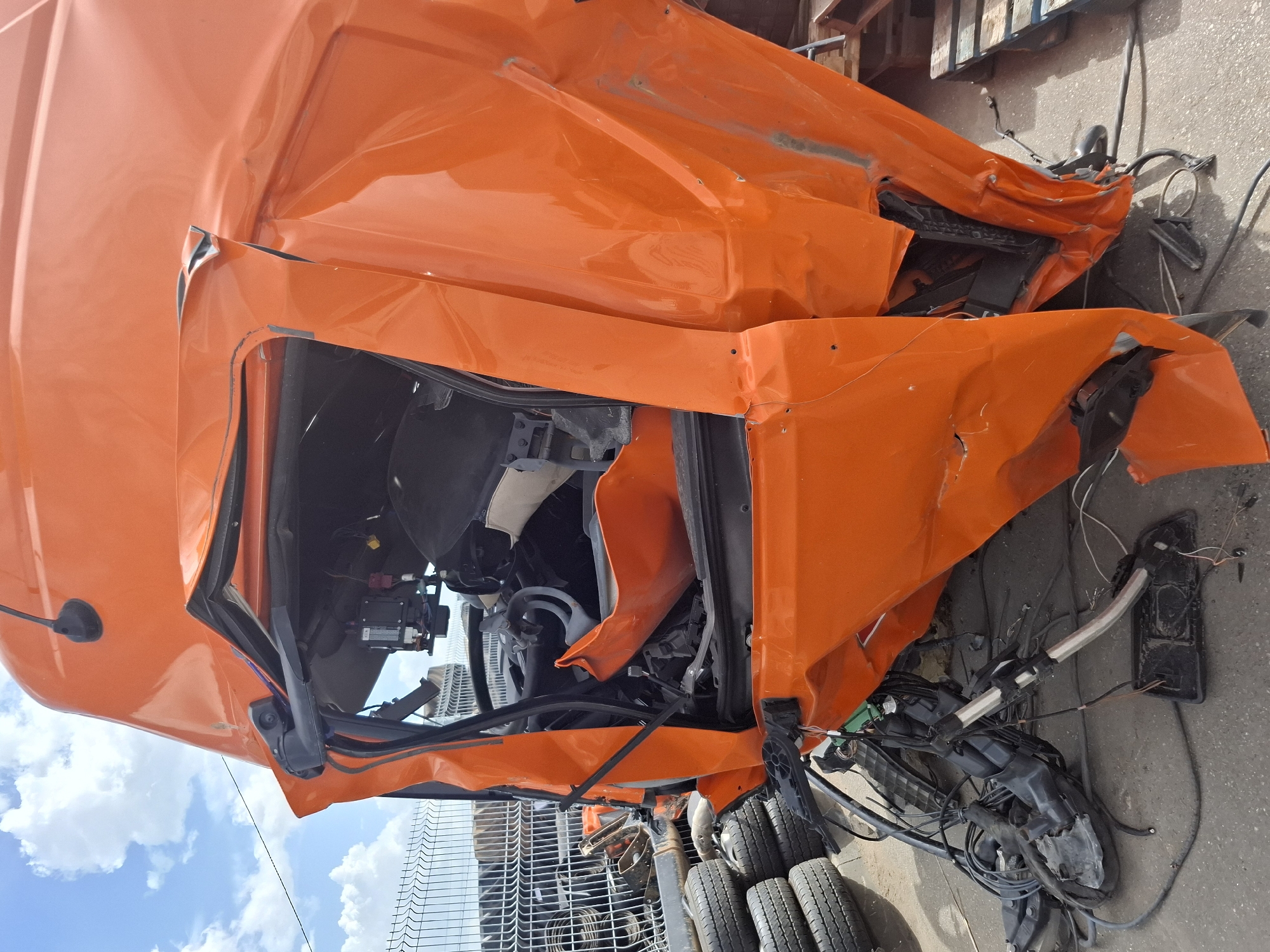 The image shows the severely damaged cab of an orange truck. The damage appears extensive, with crushed and twisted metal evident throughout the cab structure. The interior of the cab is exposed, revealing a mangled steering wheel and assorted damaged components. Numerous wires and hoses are dangling from the wreckage. The truck is located outdoors, with tires and what appears to be a metal fence visible in the background. There is evidence of pavement and possibly other debris nearby. The sky is partially visible, suggesting it is daytime. The location of this scene in Izhevsk, Russia is based on the image metadata and requires further investigation to confirm this information.
