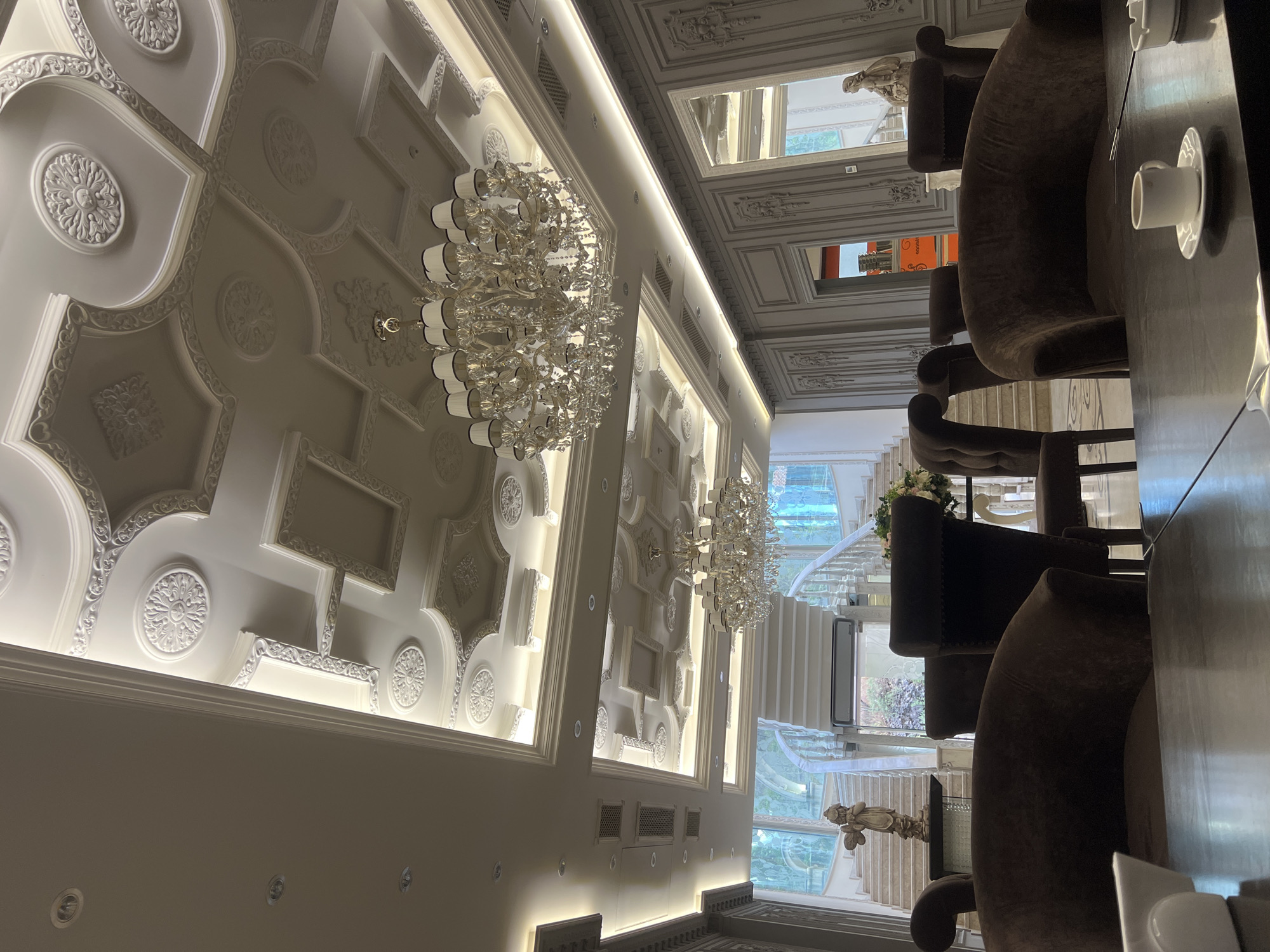 This media file captures a lavish and ornately decorated interior space, likely a restaurant, upscale cafe, or hotel lobby in Yerevan, Armenia. The scene is dominated by a highly detailed white ceiling, featuring intricate decorative moldings, recessed panels, and circular rosettes, all illuminated by concealed strip lighting and numerous recessed spotlights. Several large, multi-tiered crystal chandeliers hang prominently, casting a sparkling glow.
In the foreground, a dark, glossy table is set with white teacups and saucers. Plush, dark brown velvet armchairs with button-tufted backs and carved legs are arranged around the table, suggesting a dining or lounge area.
Through large windows or glass walls in the background, a grand white staircase with decorative balustrades is visible, along with glimpses of outdoor greenery. Reflections on these surfaces show more ornate ceilings and structural details, hinting at the expansive and luxurious design of the establishment. Decorative statuettes, including a cherubic figure, are placed on ledges or tables. A small flower arrangement is also visible on one of the tables.
Artificial lighting, primarily from the chandeliers, strip lights, and spotlights, creates a bright and opulent ambiance, suggesting an evening setting or an interior designed for constant artificial illumination. Part of a red sign with the text "GULYAS" is faintly visible through a reflection. No people are present, and no specific activity is taking place, but the setup implies readiness for patrons.