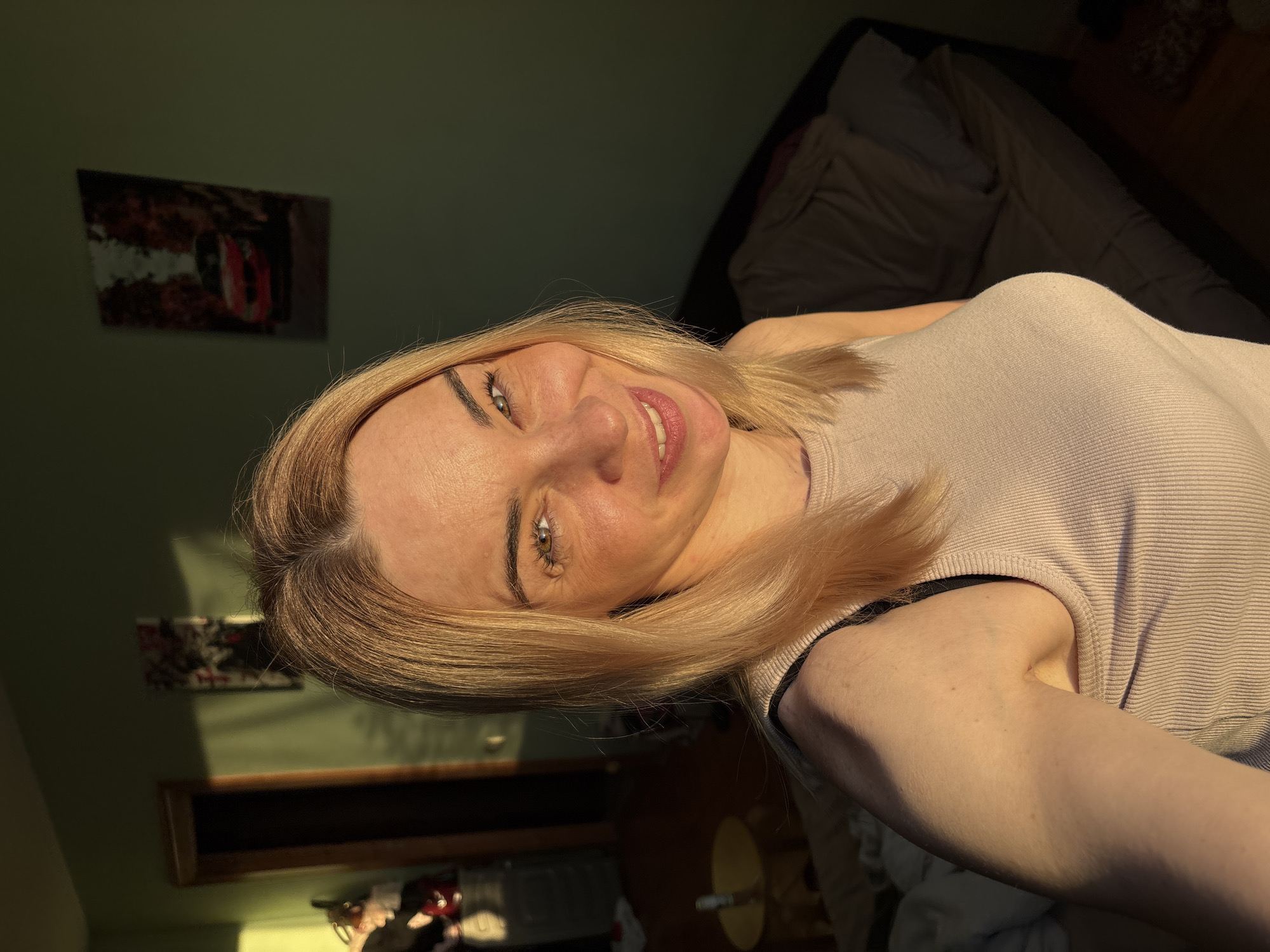 The image features a woman with light blonde, shoulder-length hair and green eyes, smiling directly at the camera. She is wearing a light, ribbed sleeveless top. Her head is tilted slightly, and she has a cheerful expression.
The setting is an indoor room, likely a bedroom or living area, with muted green walls. Strong, warm sunlight casts distinct shadows across the room and on the woman, suggesting either morning or late afternoon. Behind her, on the right side of the frame, a dark-colored bed or sofa with bedding is visible. On the left wall, a framed picture featuring abstract shapes, possibly a car or city scene in red and white tones, hangs. Below it, a wooden shelf or frame is visible. Further in the background on the lower right, a lamp with a yellow shade and other indistinct household items can be partially seen.
The woman appears to be posing for a selfie or portrait, capturing herself in the ambient sunlight. She displays a happy and friendly emotion, evidenced by her broad smile. No legible text is visible anywhere in the image.