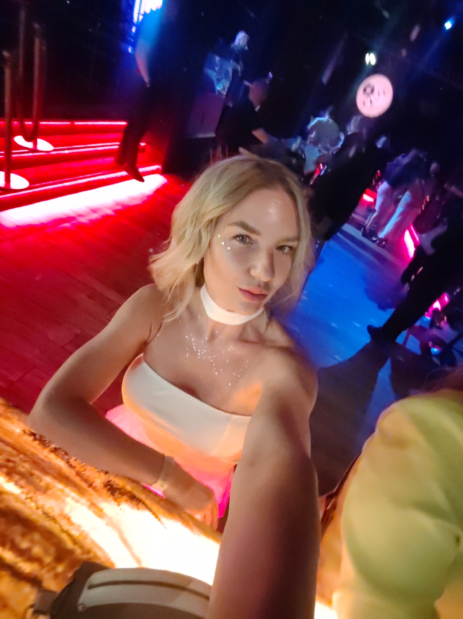 In a nightclub with red and blue lighting, a woman with blonde hair and light-colored makeup takes a selfie. She is wearing a strapless white dress and a white choker. The background shows other patrons and a DJ booth. The image is taken from a low angle, with the woman's arm and hand prominently in the foreground. The location is not specified as Petropavlovsk-Kamchatsky, Russia.
