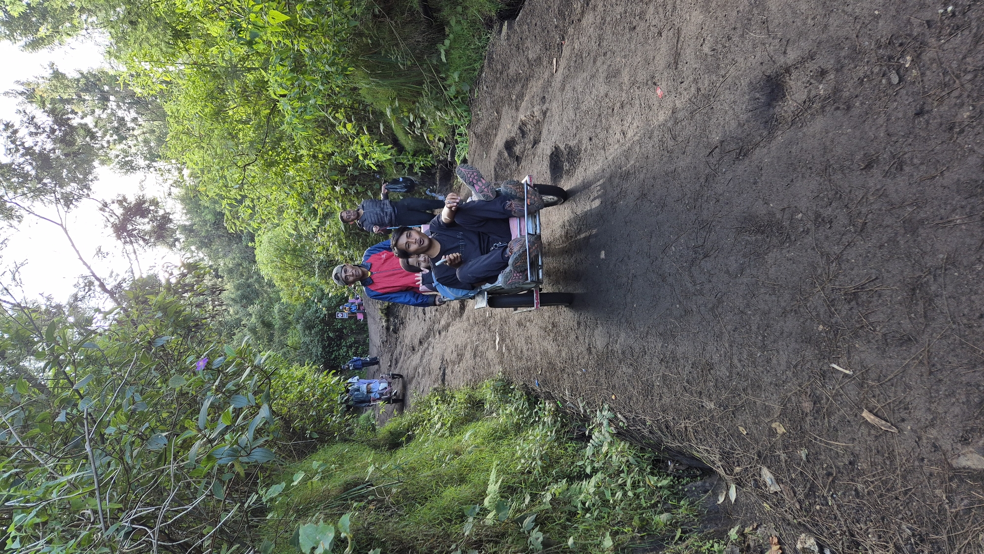 A group of people is traveling on the forest path of the city Unknown, Indonesia, riding carts. The path is muddy and lined with green vegetation on both sides.