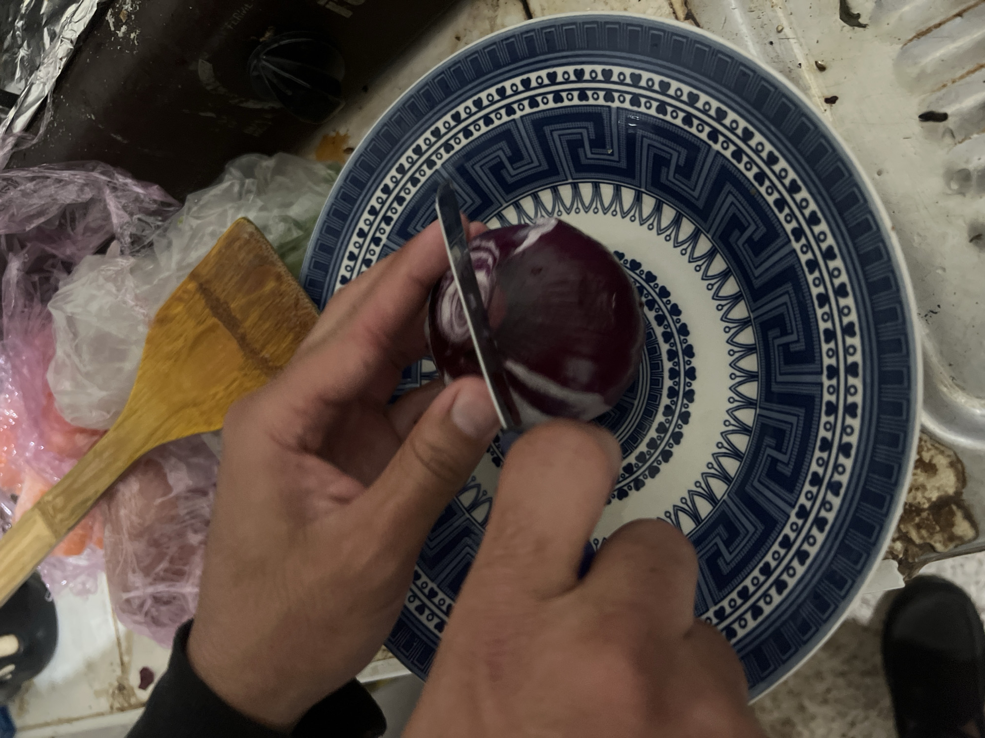 A person is cutting a red onion on a blue and white patterned plate. A wooden spatula and plastic bags are visible beside the plate. The scene appears to be indoors, likely in a kitchen. A portion of a shoe is visible in the bottom right corner of the image. The image was taken in Ouaouizert, Morocco.