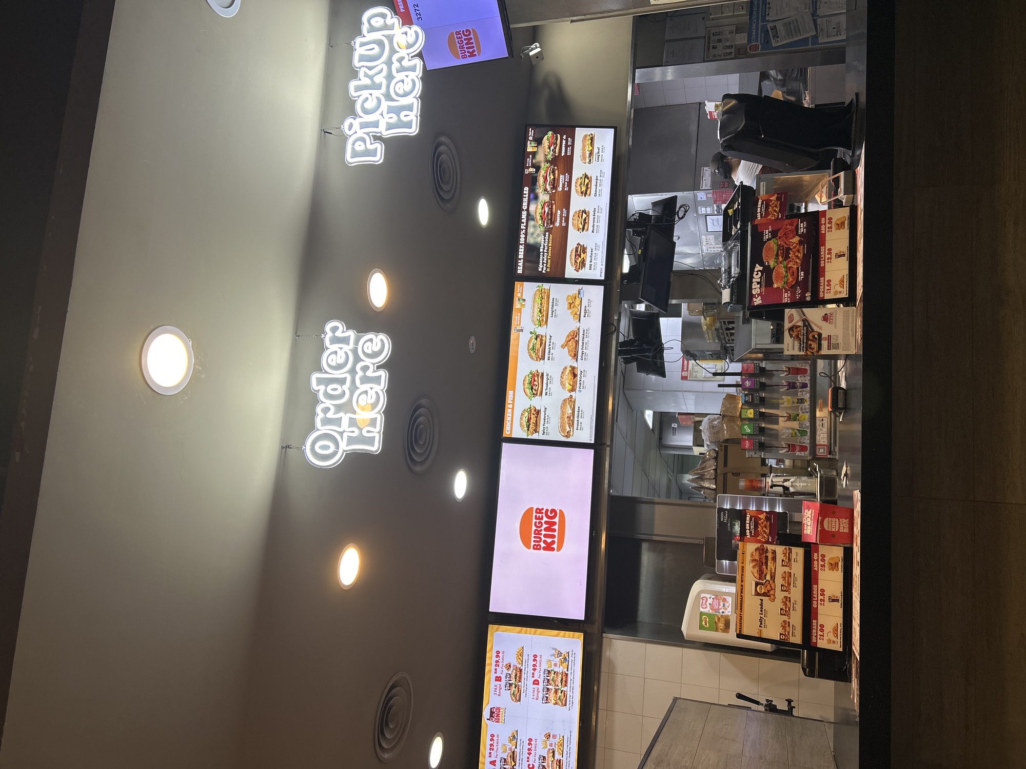 Here's a detailed description of the image:
**Location:** The image appears to be taken inside a Burger King restaurant, likely located in Subang Jaya City Council, Malaysia.
**Scene Description:**
The image showcases the order and pickup area of the fast-food restaurant. Above the counter are digital menu boards displaying various food items, prices, and promotional images. The "Order Here" and "Pick Up Here" signs are visible in bright white neon lettering. There are also ceiling lights, ventilation system components.
The counter area displays pricing for specific meal deals, including "K-Spicy". Point-of-sale systems and various condiments and supplies are visible behind the counter. It is hard to tell if there are any customers waiting.