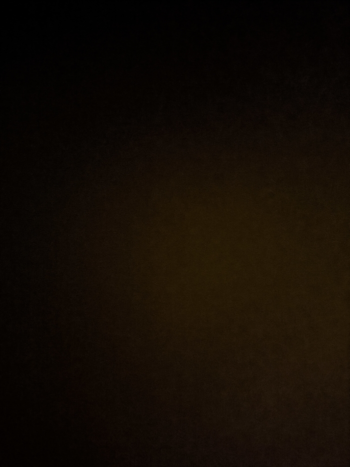 A dimly lit scene reveals a gradual gradient from near-black at the top to a dark, muted yellow at the bottom. The image has a textured appearance, possibly due to low light conditions and image noise. The color variance suggests a subtle light source originating from below the frame. The overall tone is somber, with minimal detail visible due to the darkness.