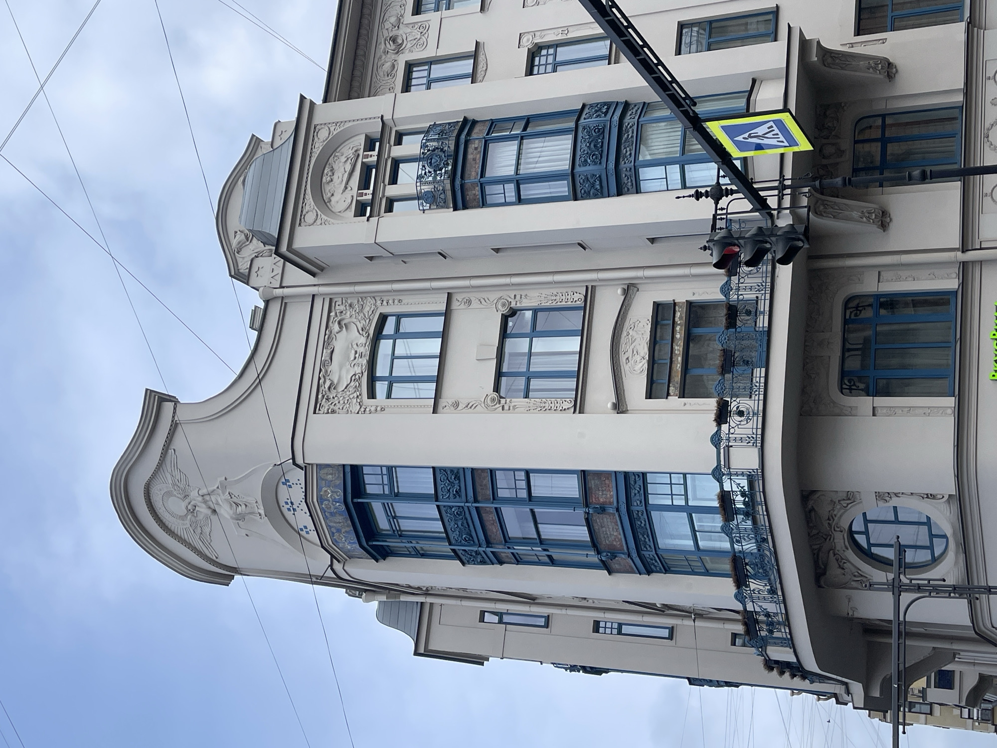 The image shows a building in Saint Petersburg, Russia. It is an ornate, multi-story structure with a white facade. The windows are framed in blue and there are decorative elements such as bas-reliefs and balconies. A pedestrian crossing sign and traffic light are visible attached to the building. Overhead wires are strung across the sky.
