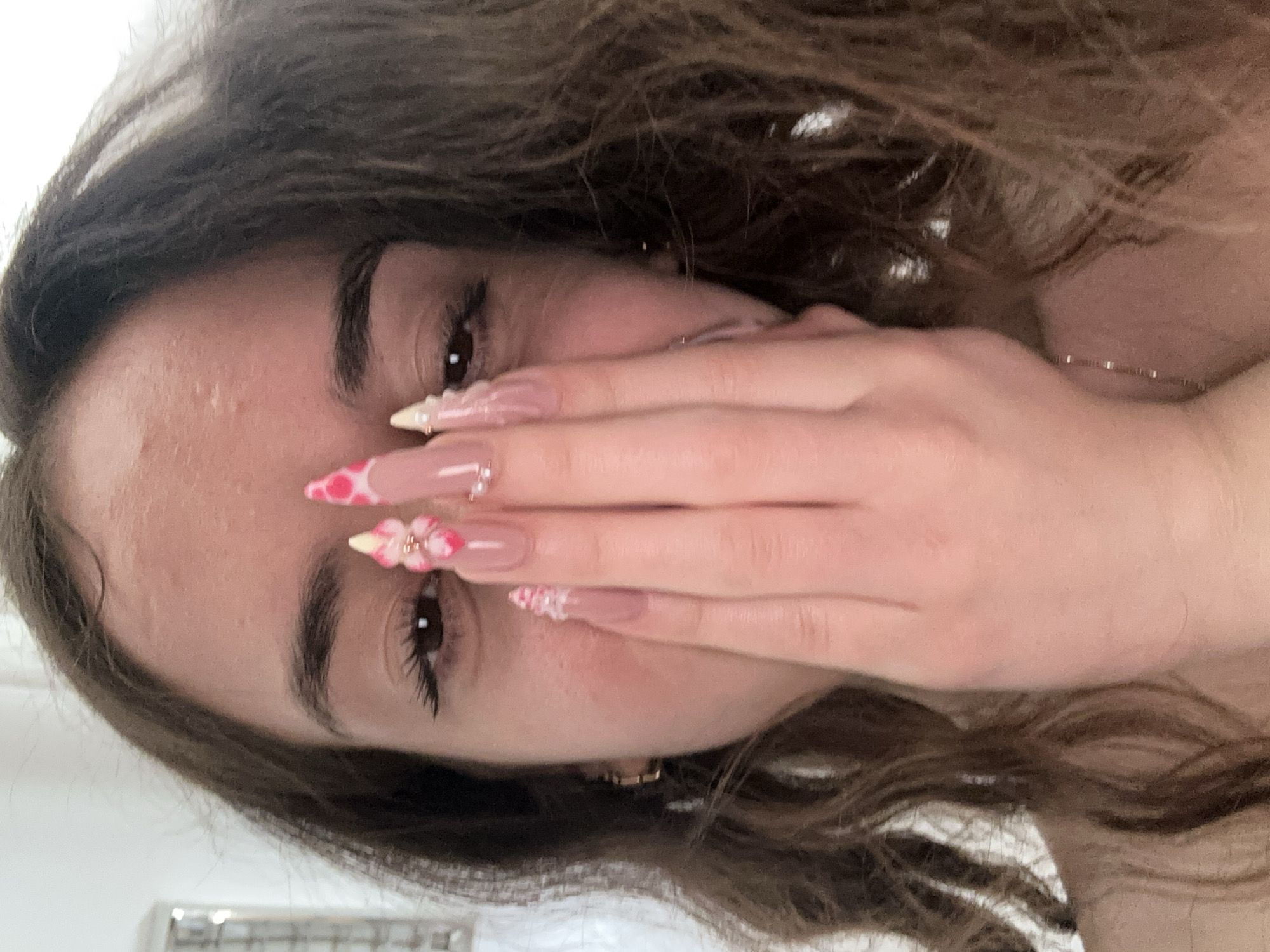 A close-up, medium shot shows a young woman from the chest up, partially obscuring her face with her right hand. Her brown, wavy hair frames her face. She is wearing a gold chain necklace. Her fingernails are long, with a clear gel polish and decorated with pink and white patterns resembling cow print, along with small floral embellishments and pearls. The background is a plain, white wall, suggesting an indoor setting. There is no visible indication of the city of Shumen, Bulgaria, in the image.