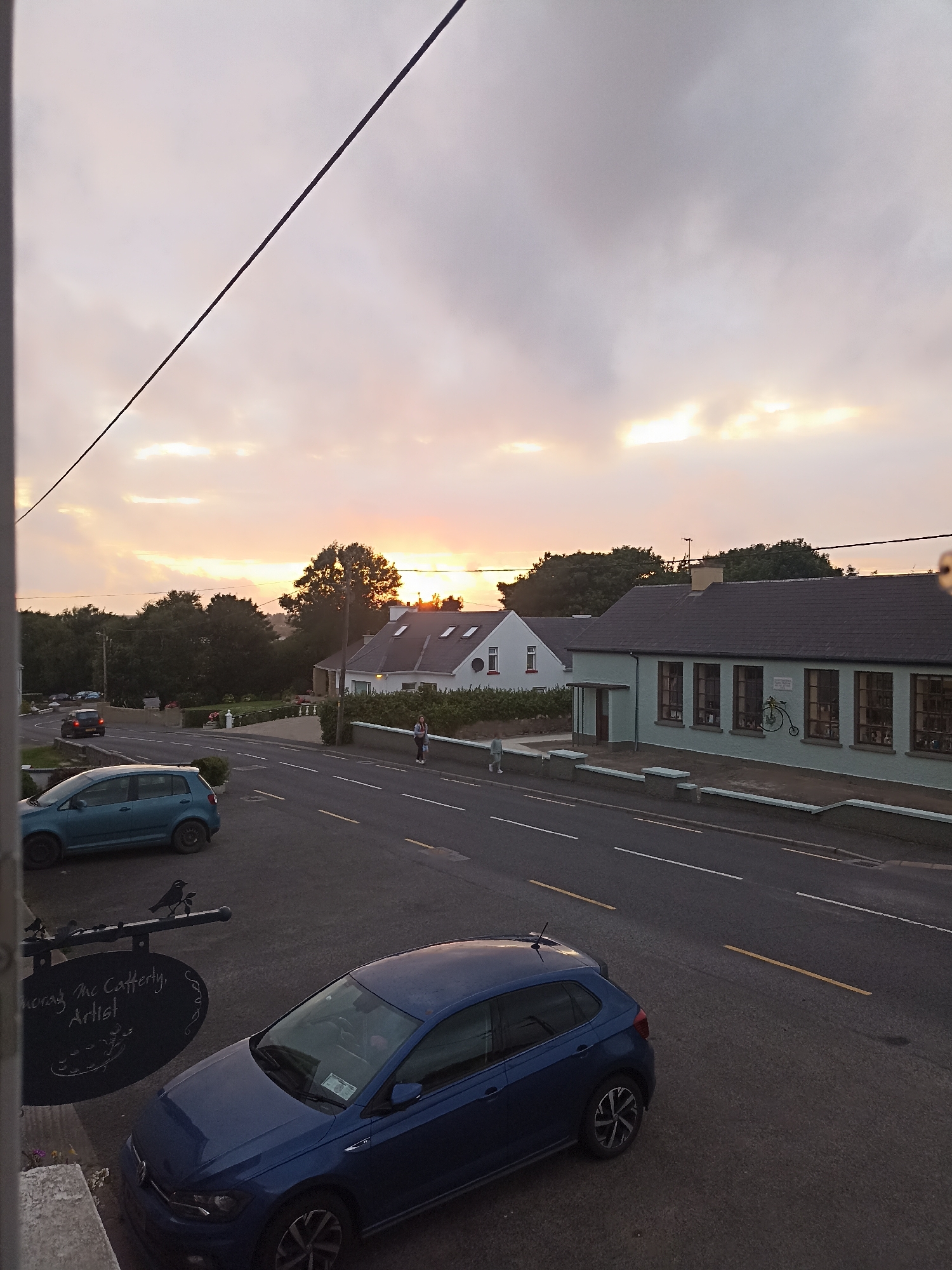 The image depicts a street scene in Annagry, Ireland, during what appears to be sunset or early dawn. The sky is overcast with a warm glow from the setting or rising sun visible in the distance, peeking through the clouds. A dark power line bisects the upper portion of the image diagonally.
In the foreground, a blue Volkswagen Polo is parked on the right side of the street. To its left, a dark, oval-shaped sign hangs from a metal pole, adorned with decorative metal birds. The sign is inscribed with "Doray Mc Cafferly, Artist." A small cluster of purple flowers is visible at the base of the pole.
Across the street, a light blue hatchback is parked. The road is asphalt with white and yellow painted lines indicating lanes.
Further down the street, on the right, is a pale green building with a dark roof. It has several large windows and a small overhang at the entrance. A decorative metal bicycle is mounted on the exterior wall of this building. To the left of this building is a white house with a dark roof, featuring a red circular detail on one of its windows. A person, appearing to be a woman wearing a light-colored top and dark pants, stands on the sidewalk in front of the green building. Another figure, possibly a child, is walking on the sidewalk in the distance.
On the left side of the street, further down, is a darker colored car driving away from the camera. More trees and vegetation are visible in the background, lining the sides of the road and forming a backdrop to the buildings. The overall impression is a quiet, rural or small-town setting.