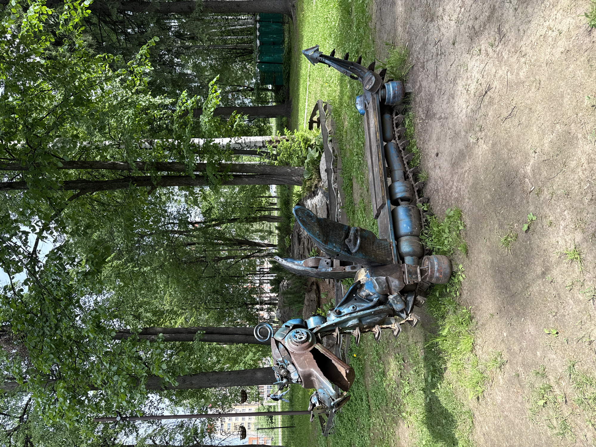 The image depicts a large, abstract metal sculpture of what appears to be a robotic dragon or mechanical beast. The sculpture is constructed from various pieces of metal, including tank treads, gears, and cut sheet metal, all painted in shades of blue and brown. It sits on a patch of green grass, with a dirt path visible to the side.
The surrounding environment is a park with trees and grass. In the background, there is a line of green portable toilets and other metal sculptures can be seen.
The sculpture is located in Izhevsk, Russia.