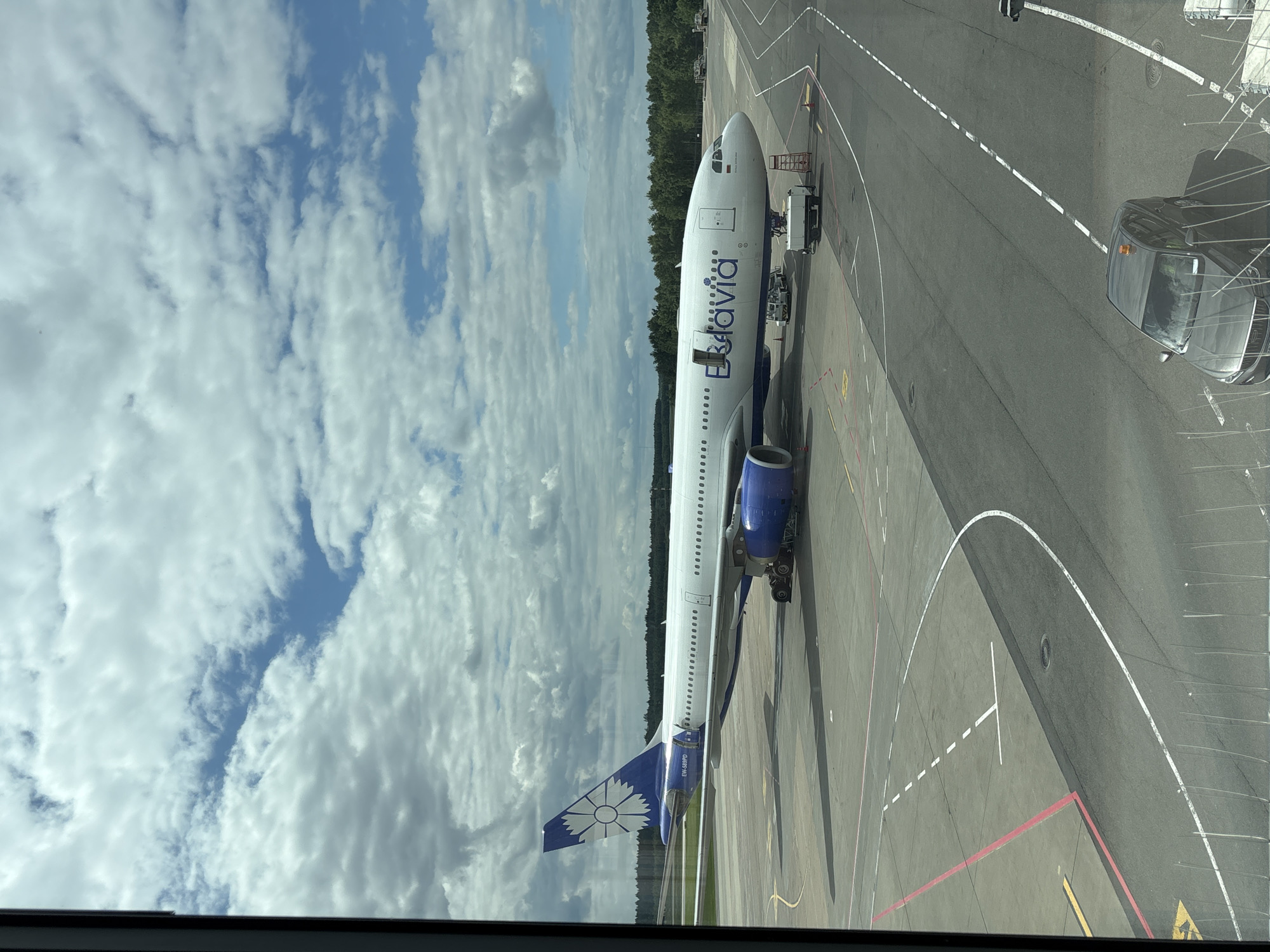 The image shows a Belavia airplane parked on an tarmac, likely at an airport in Minsk, Belarus, on a cloudy day. The airplane is white with blue accents and the Belavia logo is visible on its side. The tail of the plane features a blue design with a white flower. The tarmac is marked with various lines, including yellow and red ones, indicating taxiways and parking spots. In the background, a line of trees suggests the airport is located near a wooded area. A dark-colored car is parked on the tarmac, and other airport vehicles are visible near the plane. The scene is captured from a high vantage point, possibly from an airport terminal window. The sky is partly cloudy, with patches of blue sky visible between the clouds, indicating it is daytime. There is no clear indication of any specific activity or event taking place, other than the plane being parked. The text "EW-588PD" is visible on the tail of the airplane.