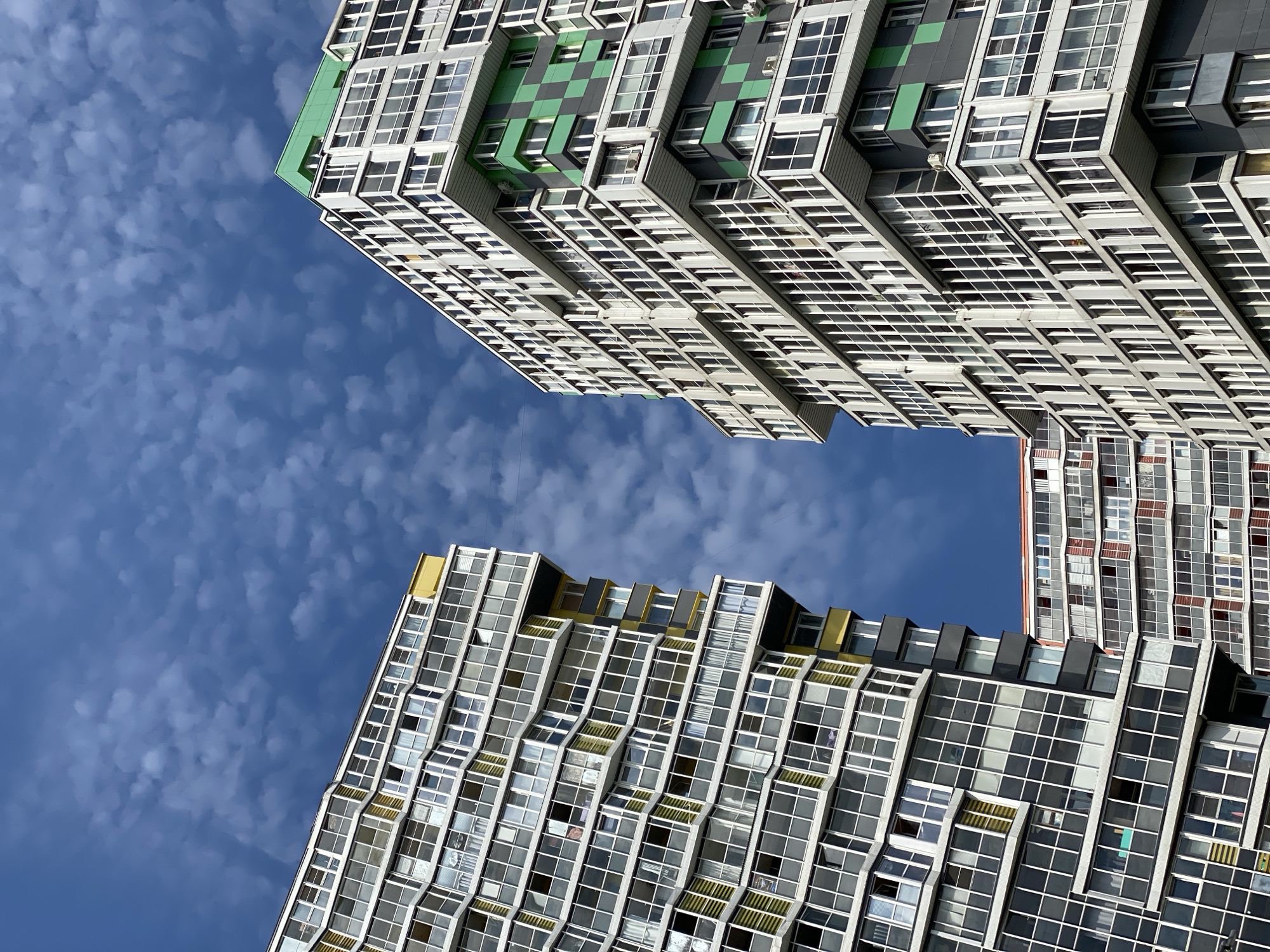The image shows a low-angle view of multiple modern residential buildings against a cloudy blue sky. The buildings are tall with numerous windows and balconies. One building on the left features yellow accents and a distinctive wavy facade. The building on the right has green checkered panels and a prominent green roof. A smaller building with red accents is visible behind the two main structures. The scene is characteristic of urban residential architecture found in Kudrovo, Russia, a developing district known for its contemporary apartment complexes. No people are visible in the image.