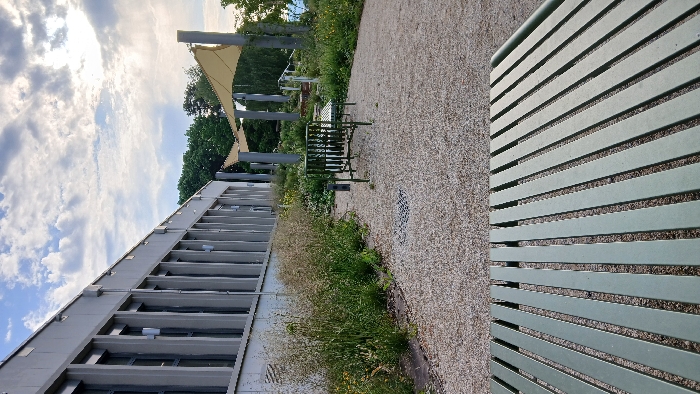 The image shows a long, gray building with many windows, located in Besançon, France at coordinates 47.246, 5.987. In front of the building is a grassy area with a gravel path. There are two metal benches positioned along the path, with a third empty bench in the foreground. The metal benches are green in color and have a slatted design. The sky is partly cloudy, and a large, yellow, fabric canopy is visible in the background. Trees and foliage are also present, with the canopy partially shading the area. The image is taken from a low angle, looking towards the building and benches. There are no people present in the scene.